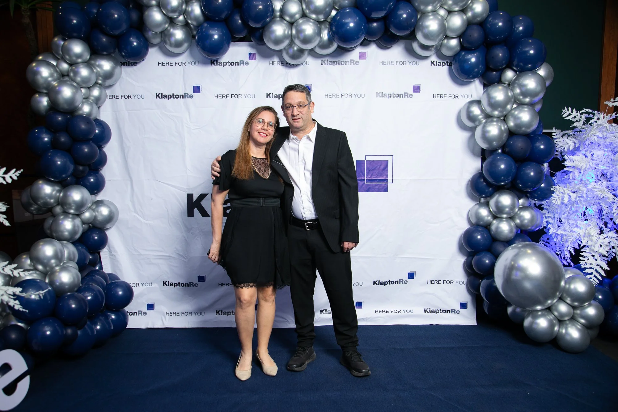 A woman in a black dress and a man in a business suit standing together, smiling, at a celebration event with a blue and silver balloon arch and a white backdrop with the text 'KlaptonRe' and 'HERE FOR YOU'.