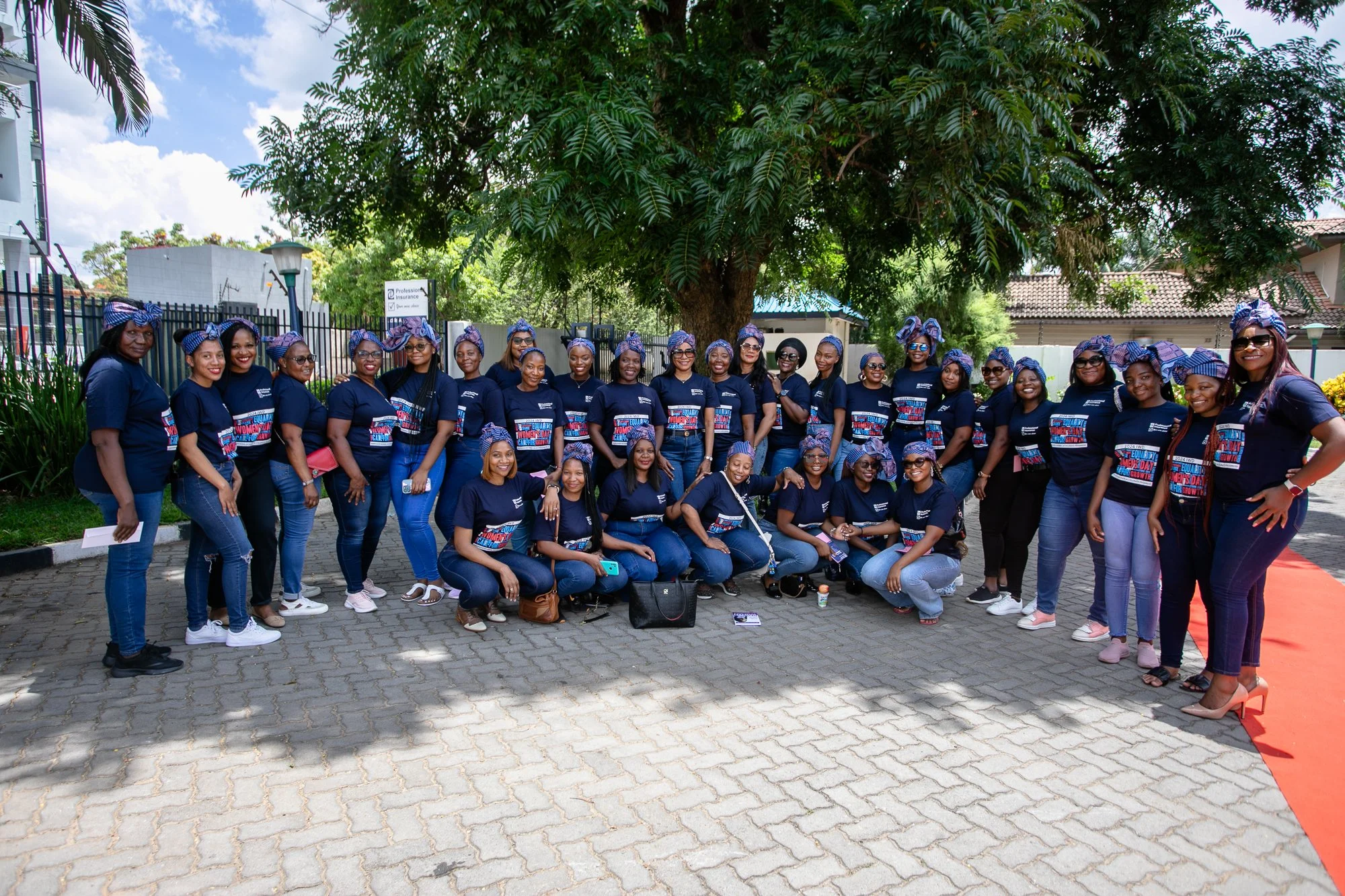 A group of women wearing matching navy-blue t-shirts and headbands, standing outdoors under a large tree, smiling for the camera. Some are standing, while others are kneeling or crouching in front, with a red carpet visible on the right side of the i