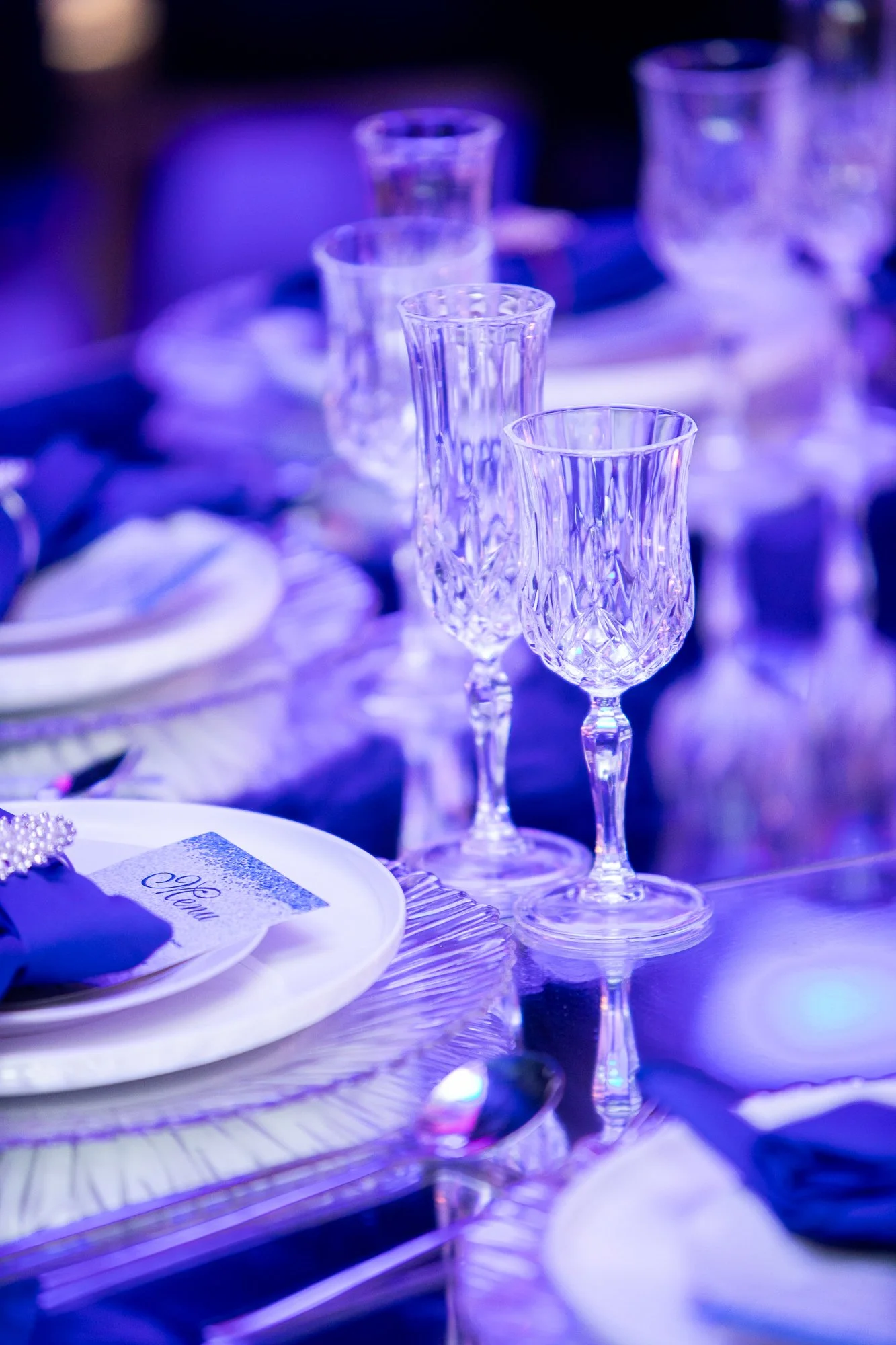 Elegant table setting with crystal glasses, a plate with a napkin, and a menu card, illuminated by blue lighting.