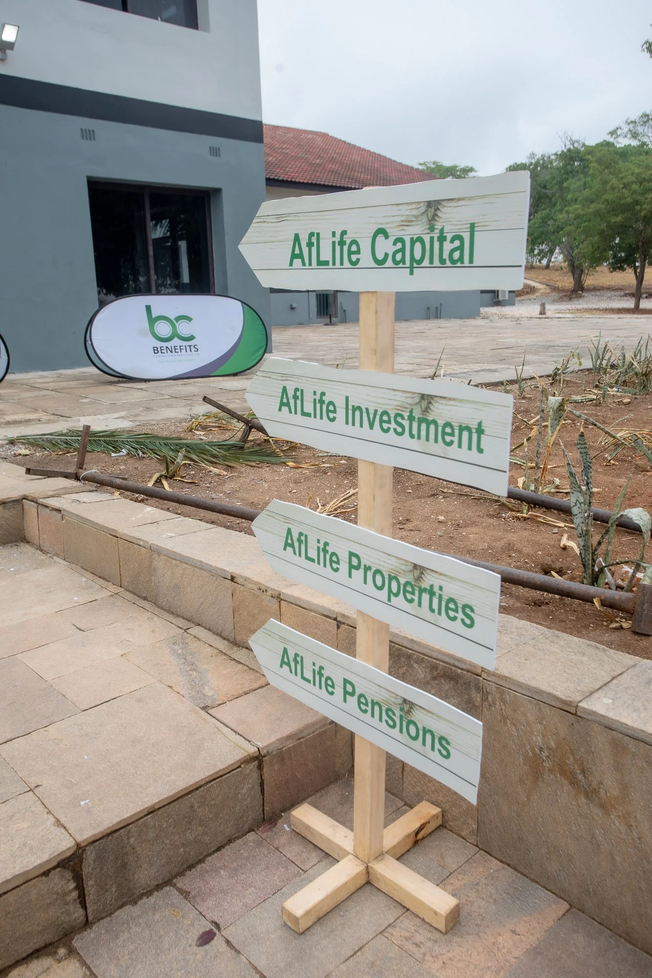 Wooden directional sign with green text pointing to AFlife Capital, AFlife Investment, AFlife Properties, and AFlife Pensions outside near a building and some trees.