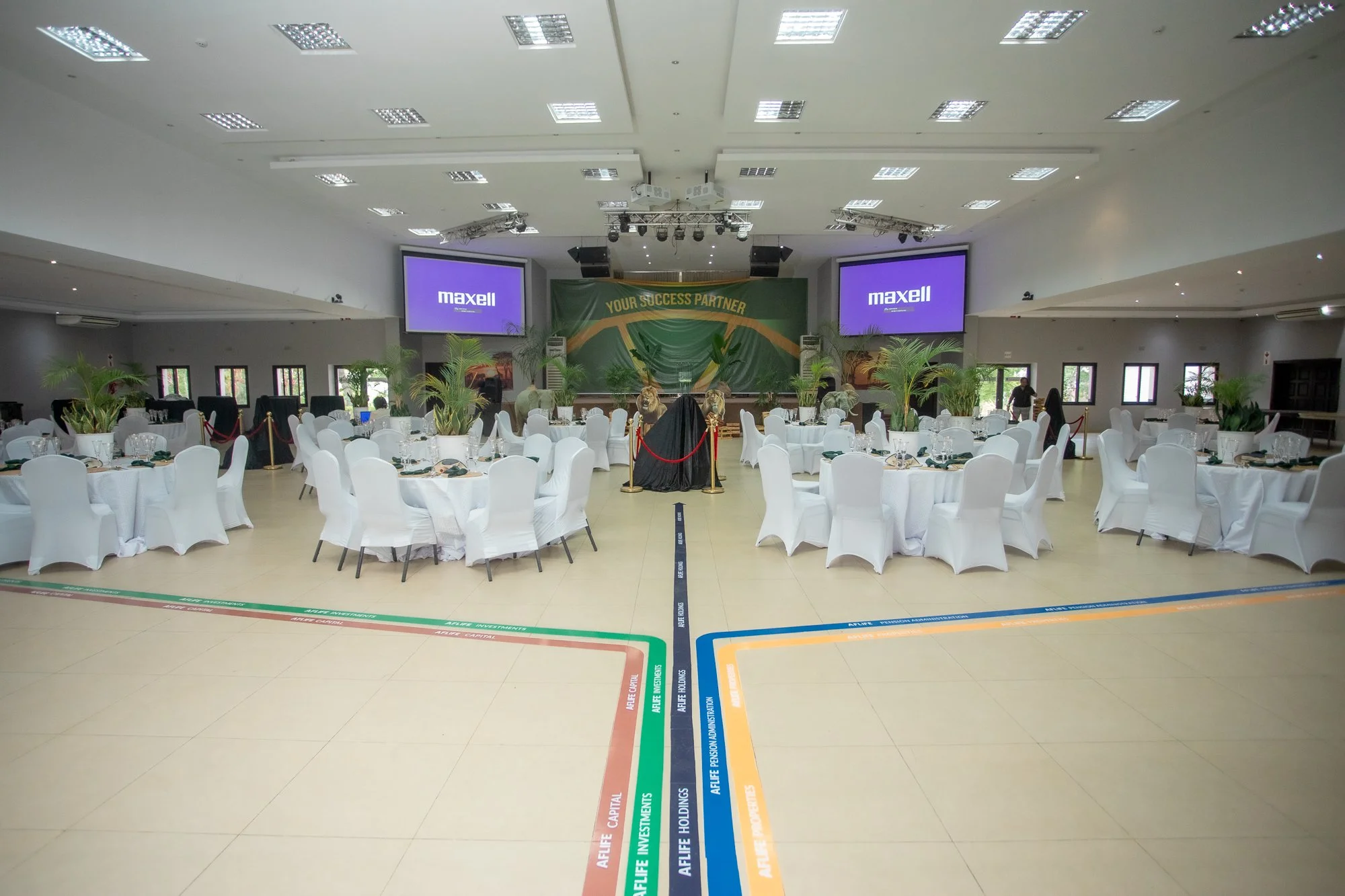 A large conference room decorated for an event with round tables covered with white tablecloths and chairs, a stage with two large screens displaying 'maxell', potted plants, and a backdrop with a slogan 'Your Success Partner'.