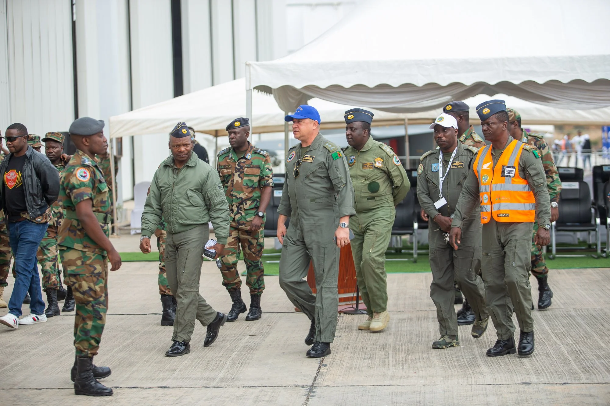 Group of military personnel walking and standing outdoors near white tents, with some individuals wearing uniforms and others in civilian clothing.