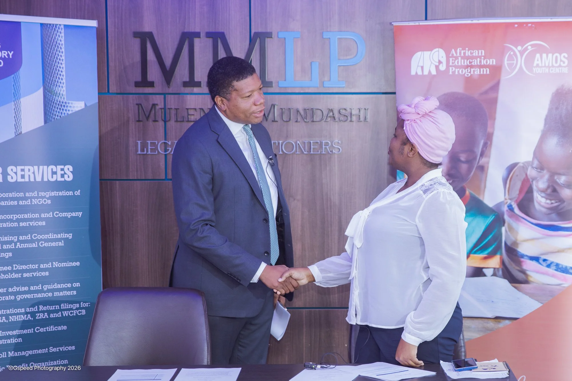 A man in a suit and a woman in a white blouse shake hands at a professional event. Behind them is a wooden wall with the words 'MULEN MUNDASHI' and 'LEGISLATORS' on it and a banner displaying logos for the African Education Program and the AMOS Youth