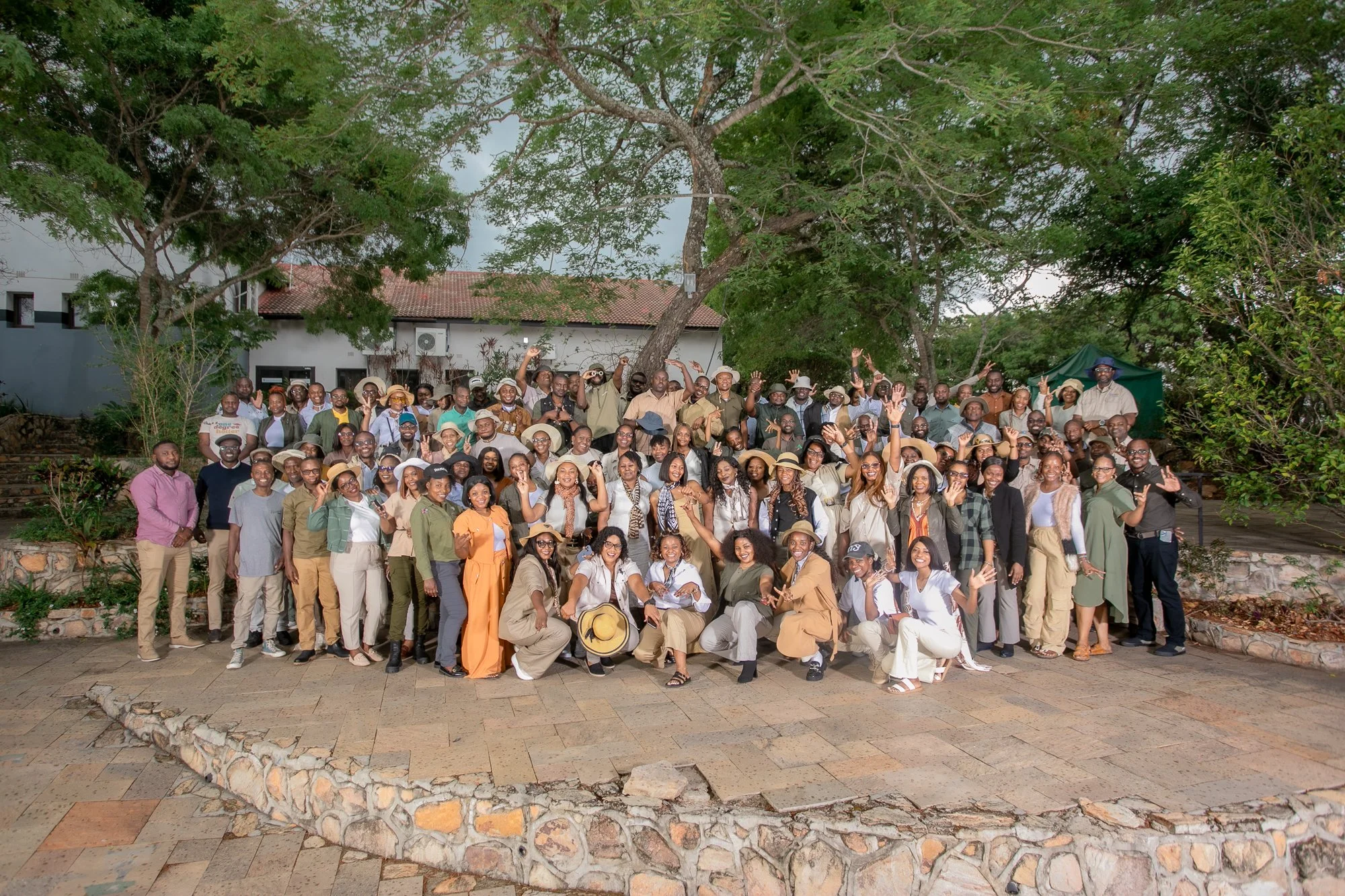 A large group of people gathered outdoors, posing for a photo in front of a building with trees around them. The group is smiling and waving, with some wearing hats and casual attire.