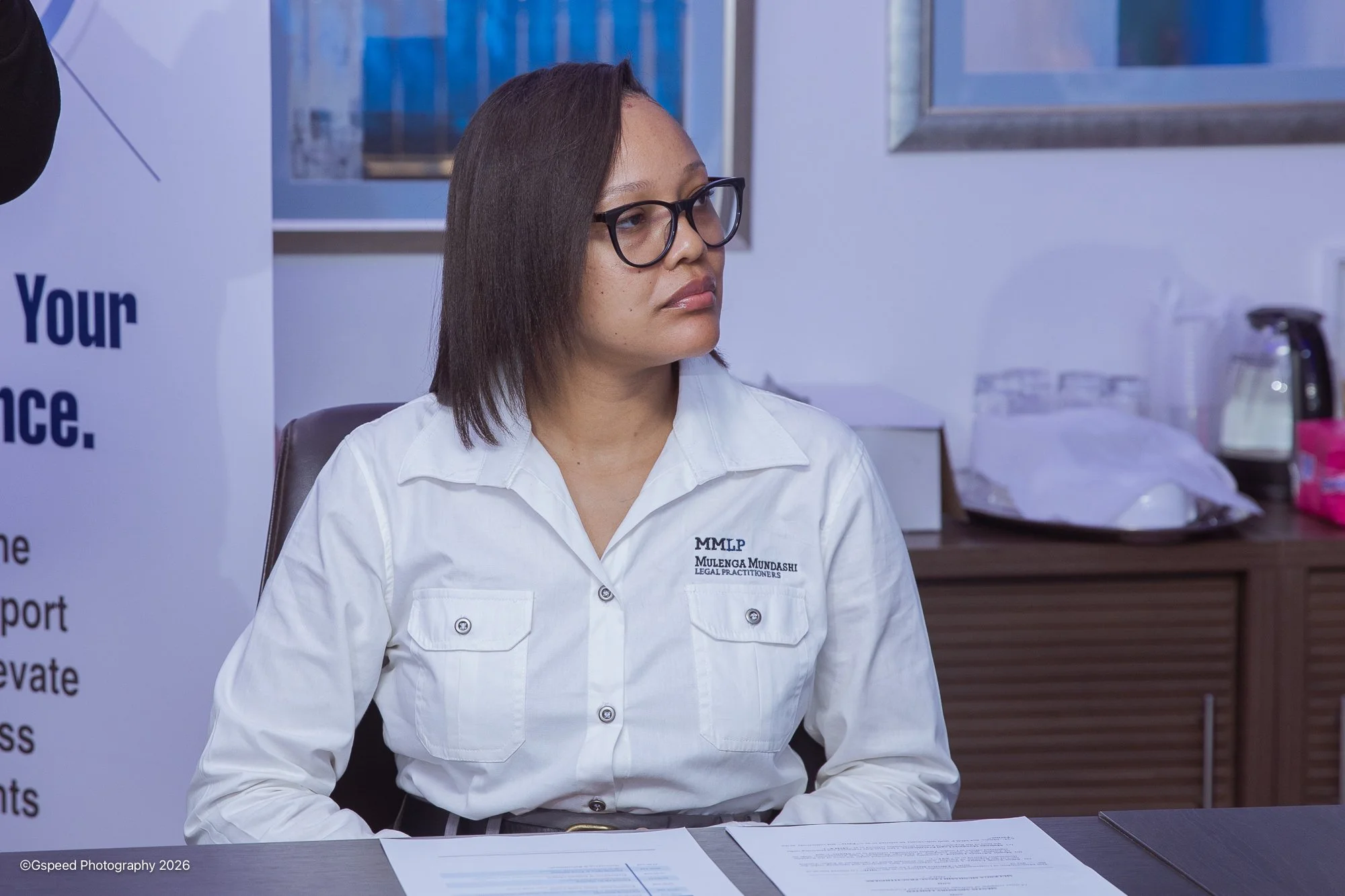 A woman wearing glasses and a white shirt with her name embroidered on it, sitting at a desk with papers, in an office or medical setting.