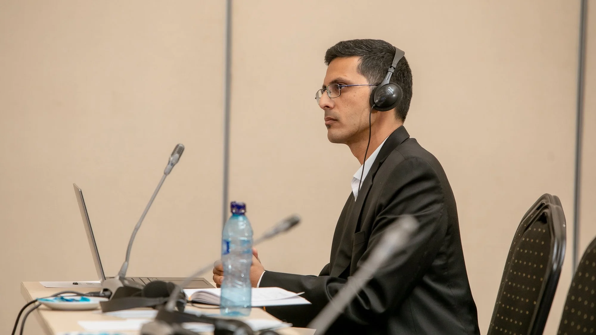 A man wearing a black suit, glasses, and a headset sitting at a conference table with a laptop, microphone, and water bottle during a meeting or presentation.