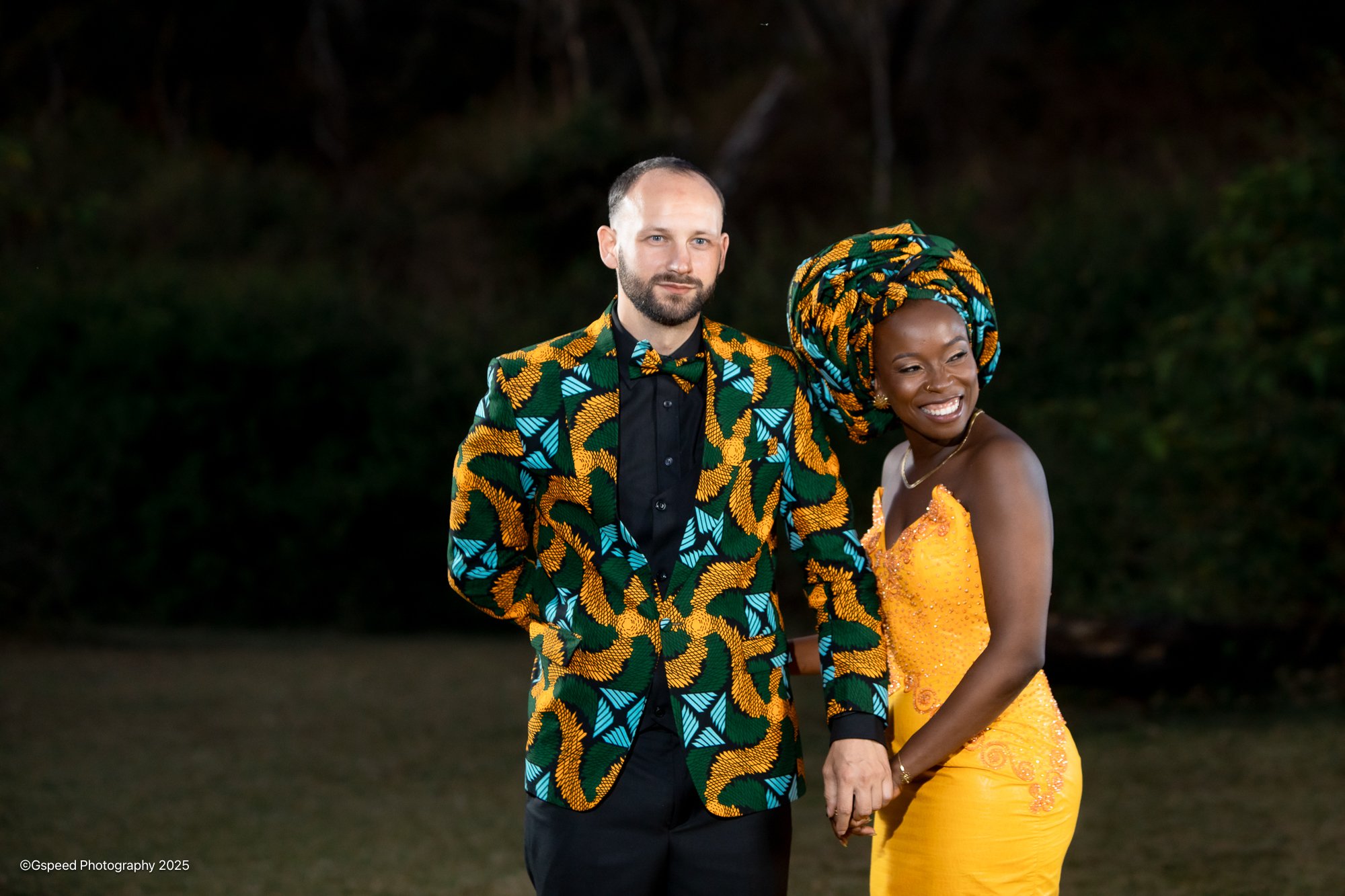 A couple dressed in vibrant, colorful African print attire, holding hands outdoors at night, with a dark background of trees.