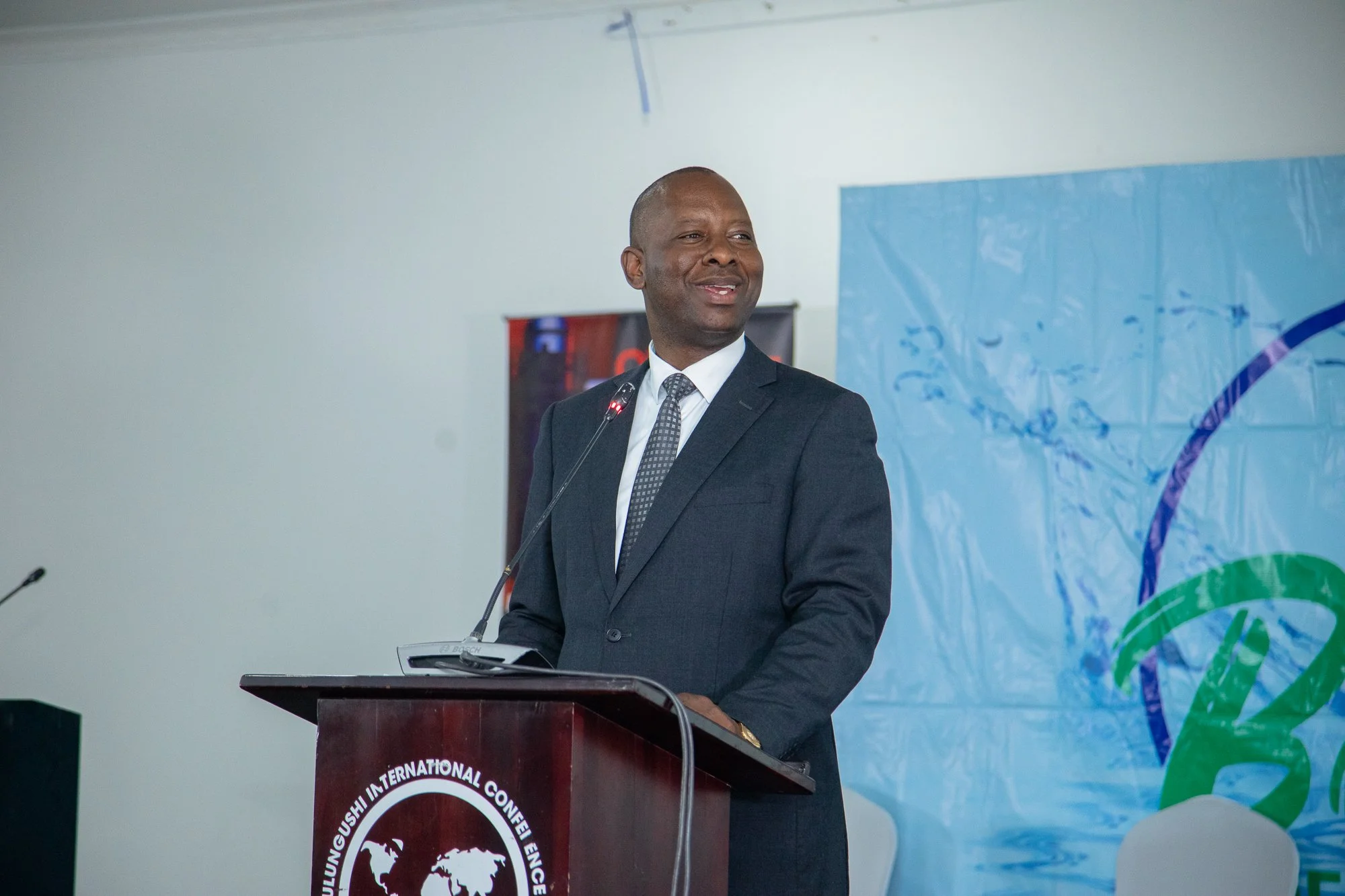 A man in a dark suit and tie stands at a podium delivering a speech at an event. There is a blue banner and a globe graphic behind him.