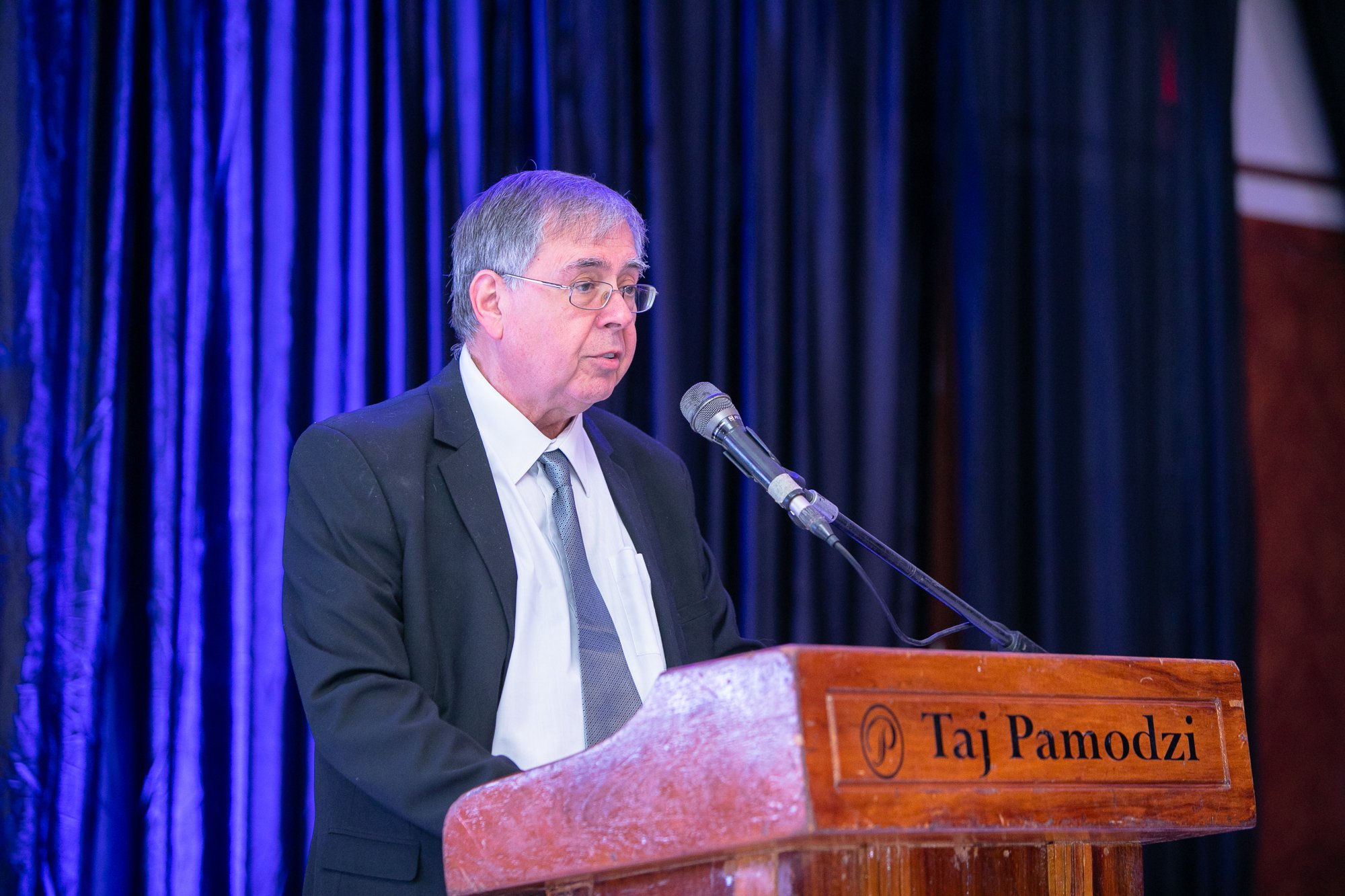 A man with gray hair, wearing glasses and a dark suit, stands at a podium speaking into a microphone at an event. The podium has a sign with the name Taj Pamodzi. The background features blue curtains.