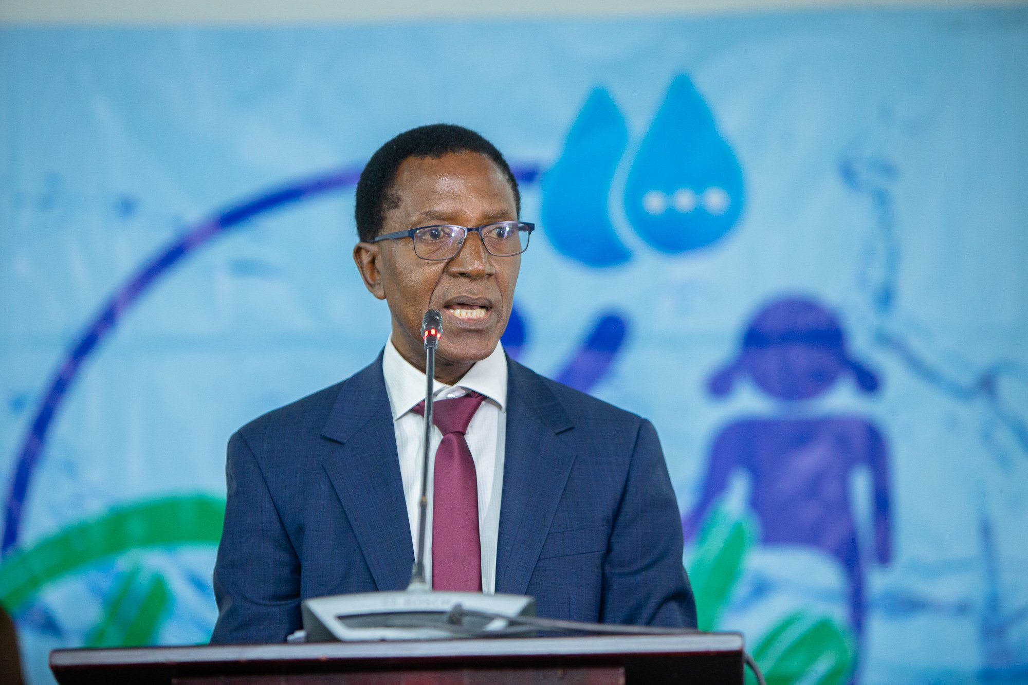 Man in a suit and glasses speaking at a podium in front of a blue branded backdrop with water droplet graphics.