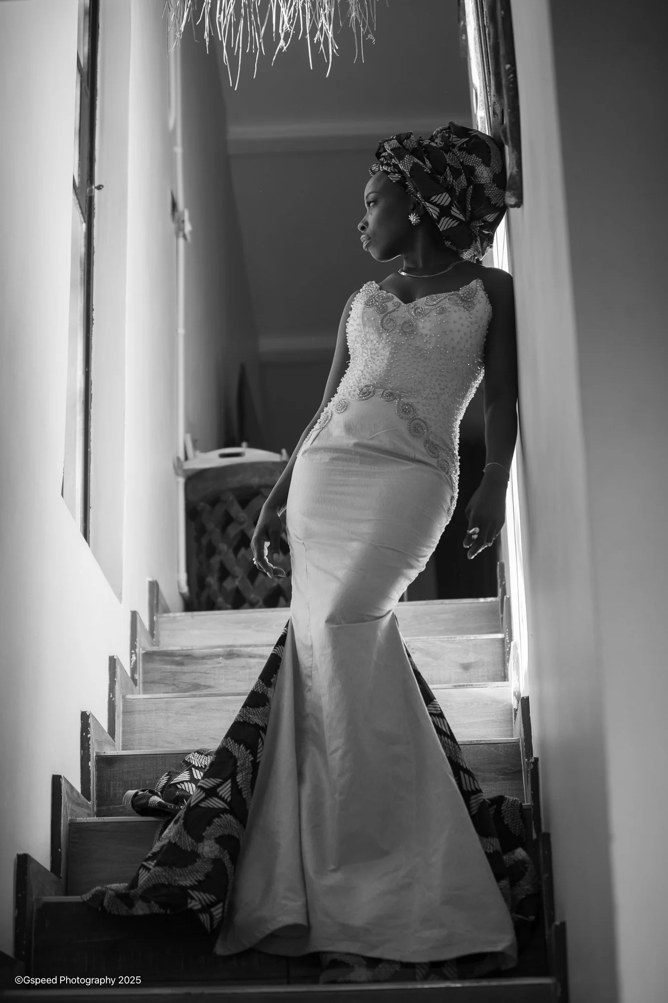 Black and white photograph of a woman in a wedding dress standing on a staircase, looking to her right, with her head wrapped in a patterned headscarf.