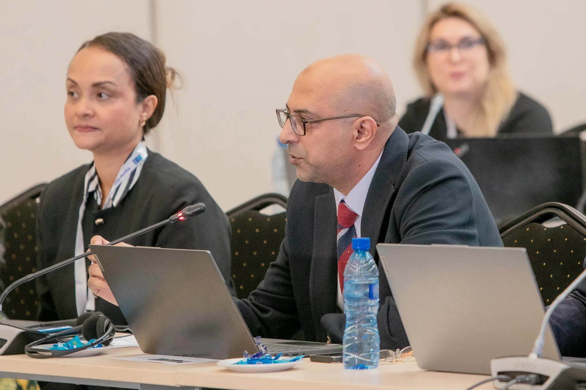A group of three people in a conference room, sitting at a table with laptops. The man in the foreground is bald, wearing glasses, a black blazer, a white shirt, and a red tie with blue designs. He has a water bottle in front of him. The woman on his
