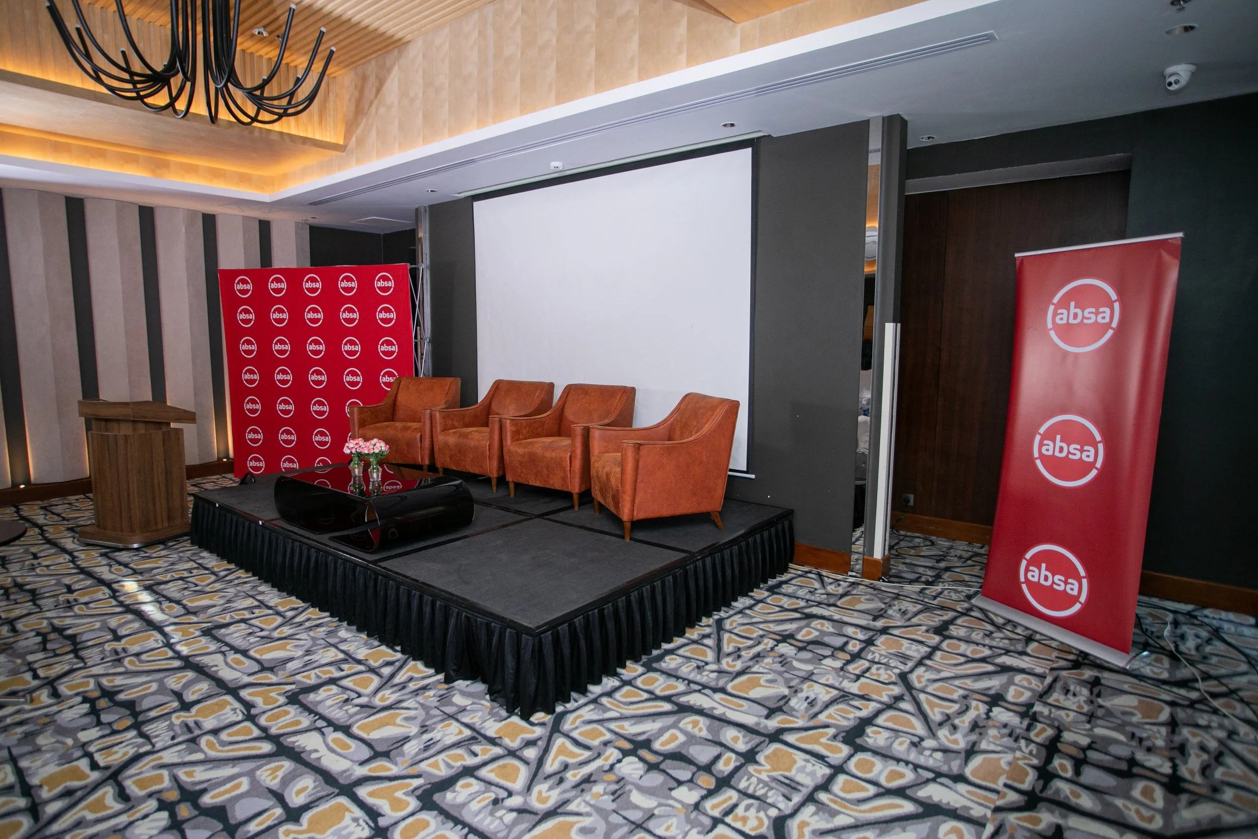 An indoor conference setup with a stage displaying a podium, three brown chairs, and a table with flowers. Red banners with the Absa logo are on either side of the stage. The room has a patterned carpet, wood-paneled walls, and ceiling lighting.