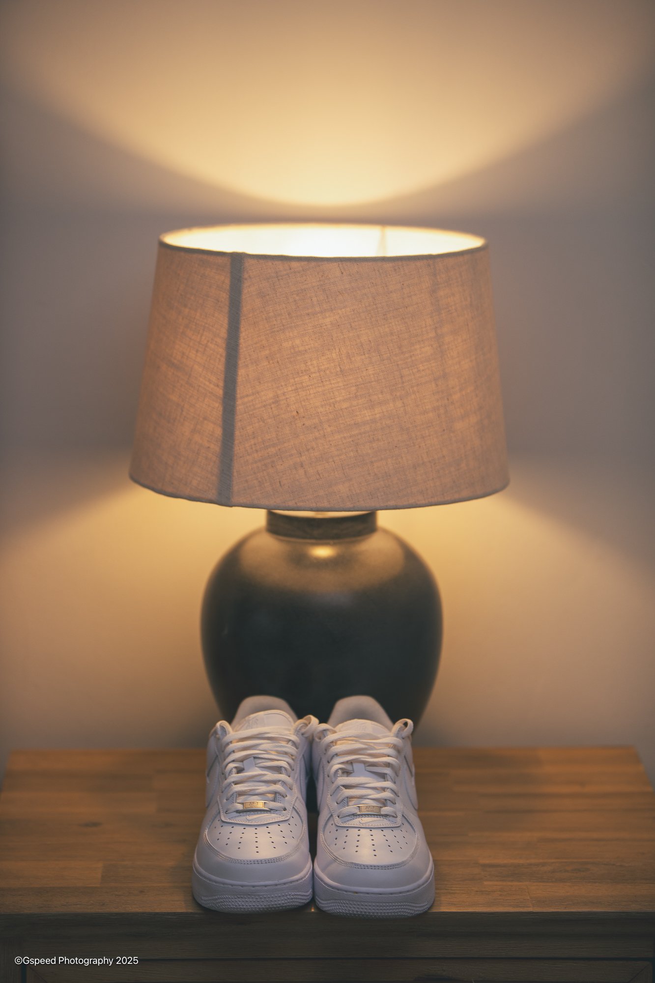 A pair of white athletic shoes, a black table lamp with a beige lampshade, and a decorative white rope knot on a wooden surface.