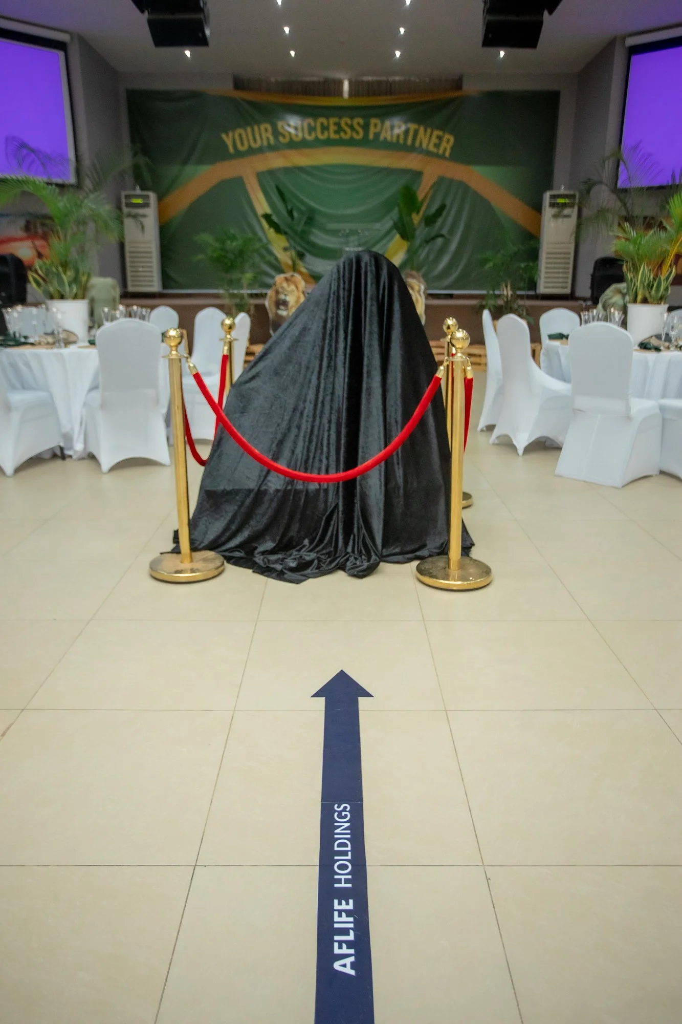 A black cloak covers something on a table with a red rope barrier in front. In the background, there is a green banner with yellow text that says "YOUR SUCCESS PARTNER," and a stage with plants and large screens on each side. The floor has a blue arr