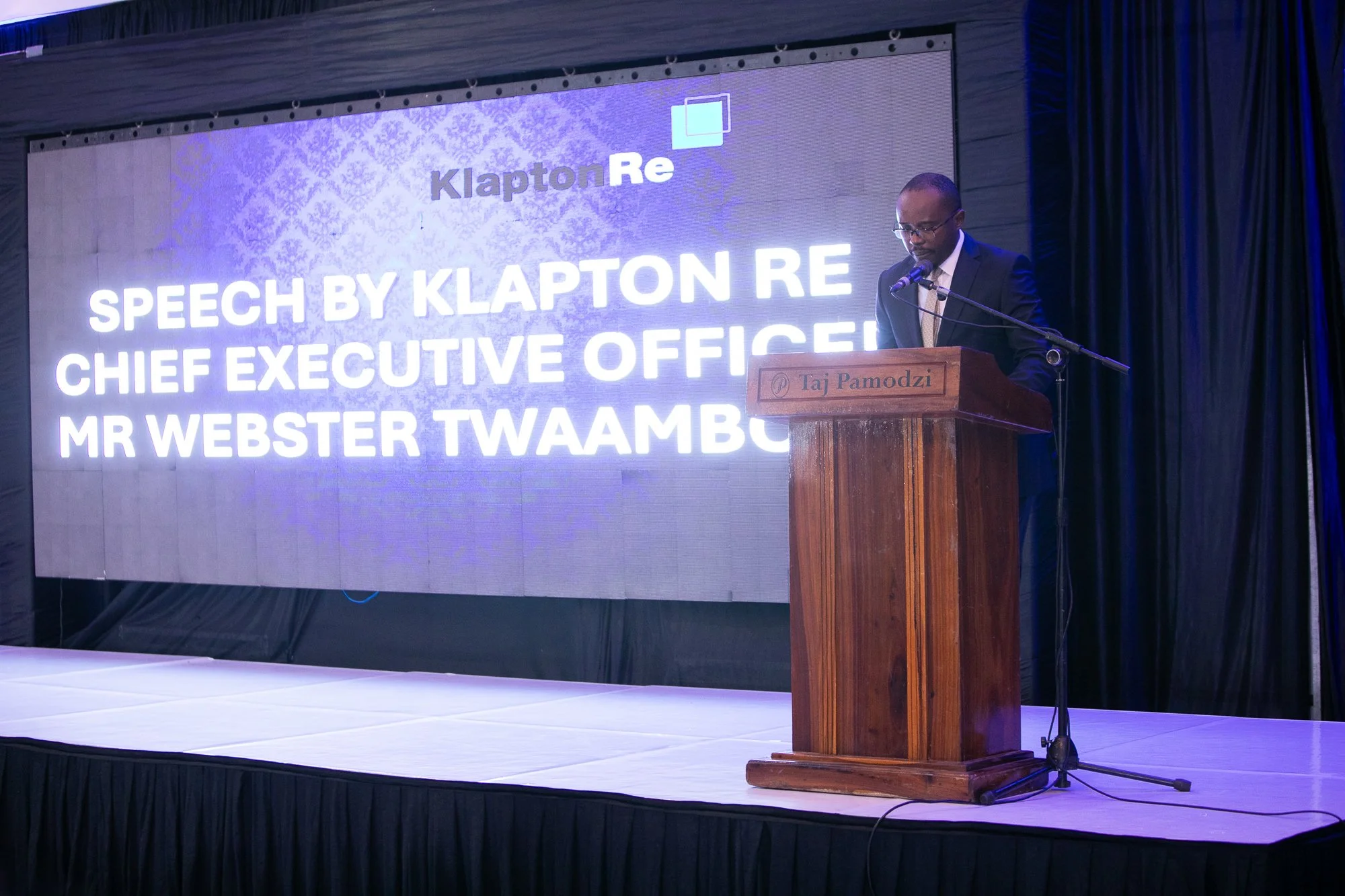 A man in a suit speaking at a wooden podium with a microphone, in front of a large digital screen displaying a presentation slide about KlaptonRe and a speech by Mr. Webster Twaambo.