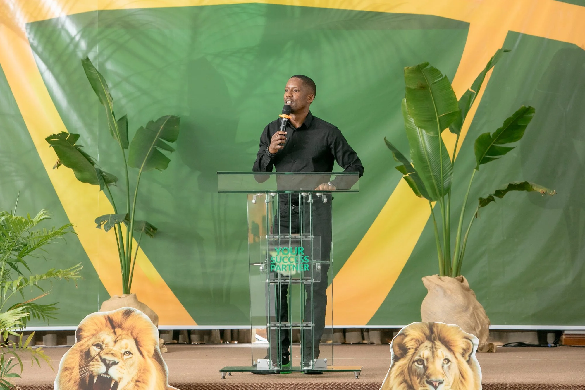 A man standing at a clear podium with a microphone, speaking or giving a presentation. The background features a large green and yellow banner with tropical plants around the stage, and cutouts of lion faces at the front of the stage.