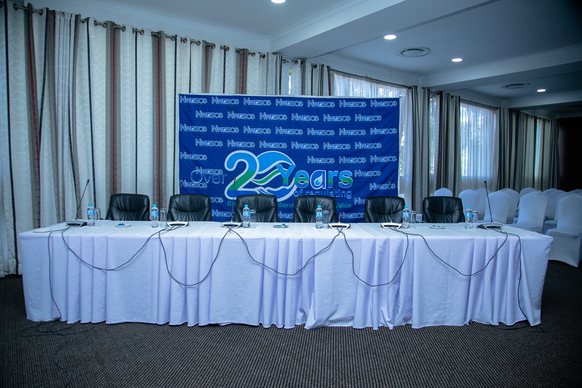 Conference room with a long white table, black chairs, water bottles, microphones, and a large blue banner that reads 'Over 20 Years of delivering value' with a logo containing 'Nwasce' repeated in the background.