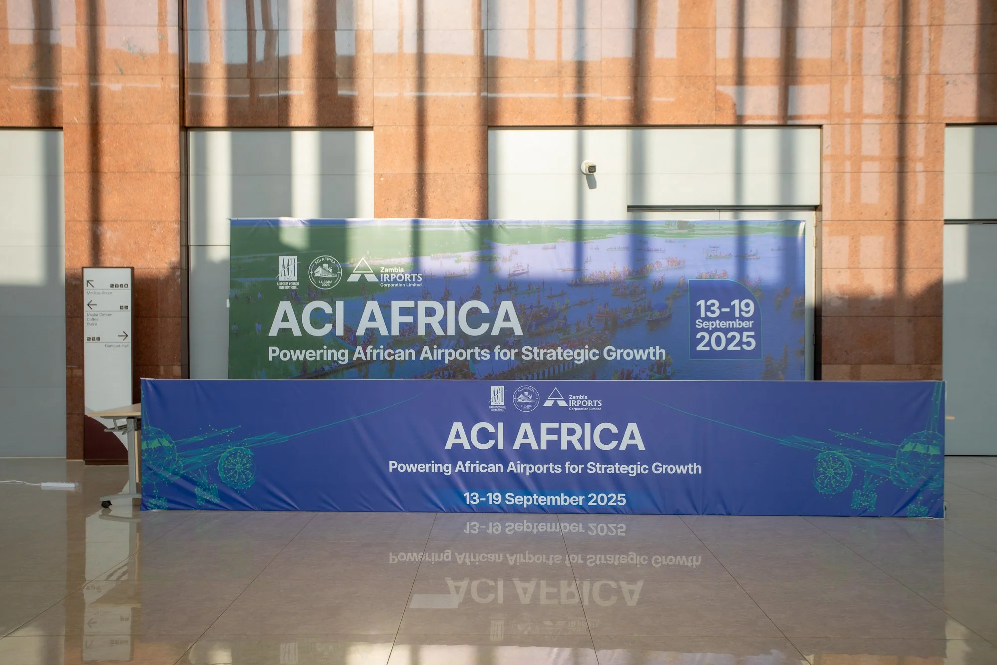 A large banner inside an airport terminal reads, "ACI AFRICA Powering African Airports for Strategic Growth 13-19 September 2025." The background shows a building with red-brown brick walls and large windows, with shadows from vertical structures cas