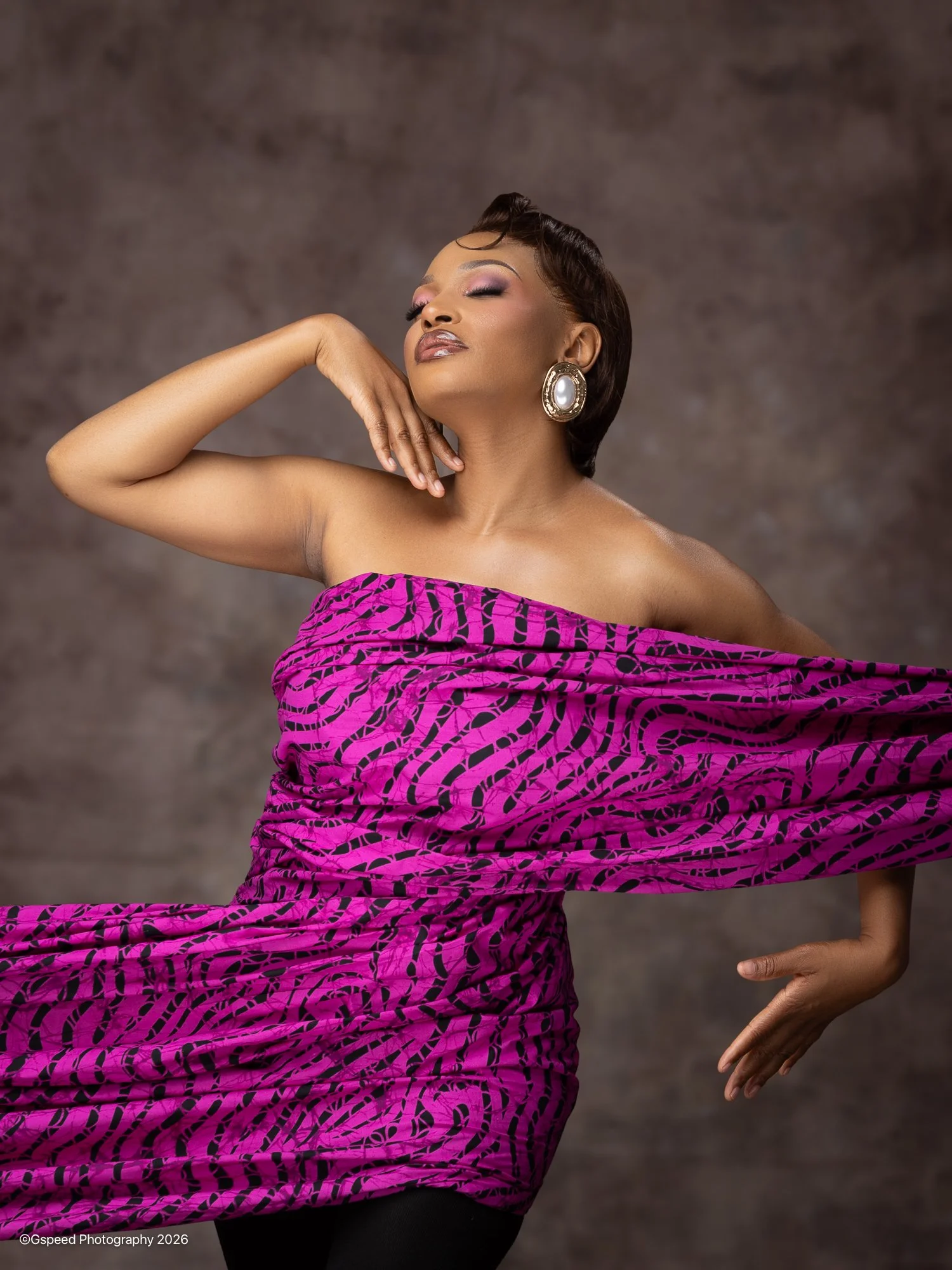 Woman with short dark hair and jewelry posing with eyes closed, wearing a pink and black patterned wrap dress, against a gray background.