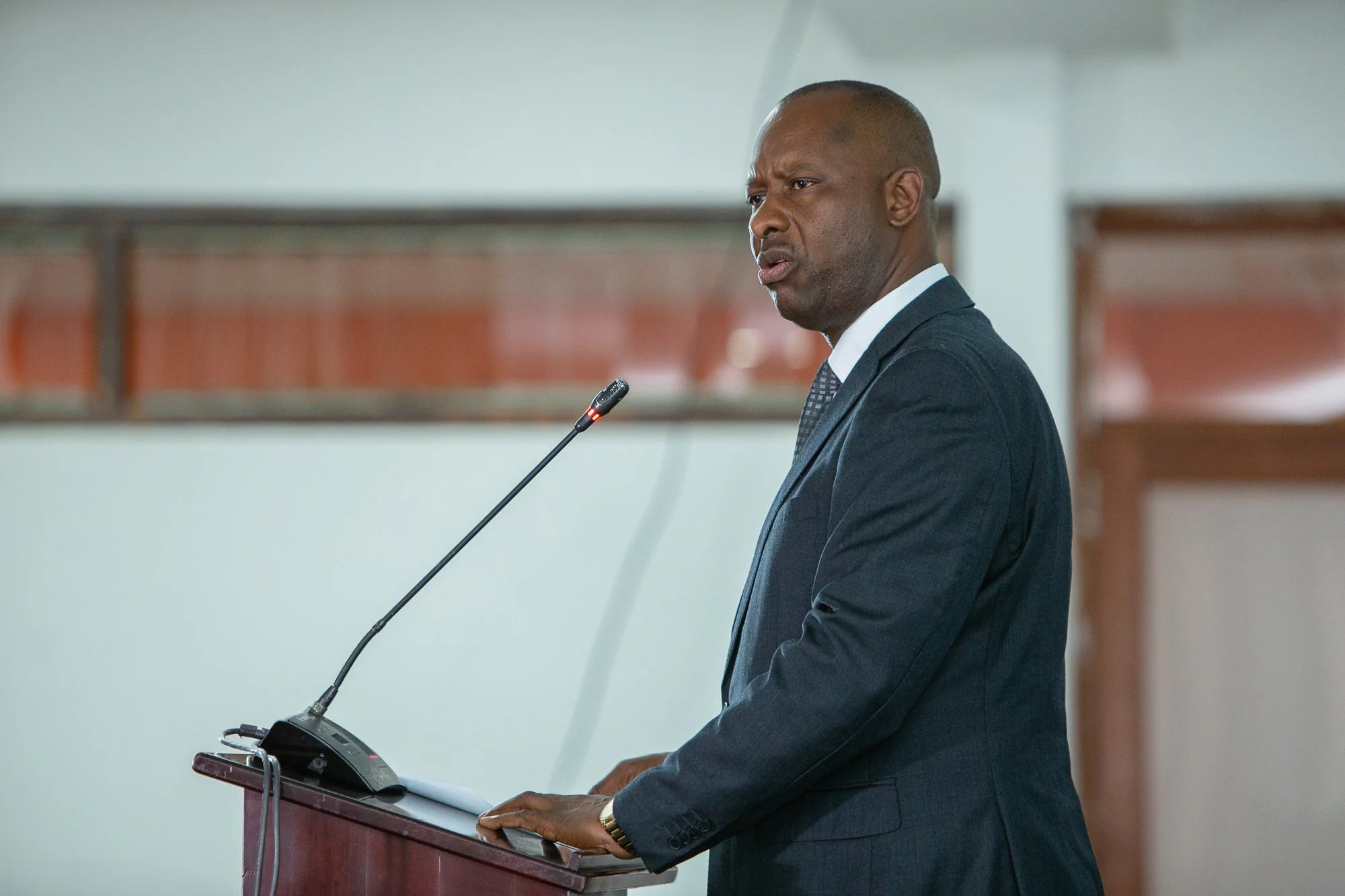 Man in a navy suit giving a speech at a podium with a microphone in a large room.