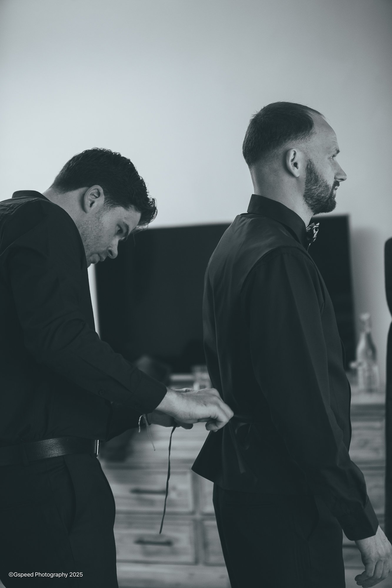 Two men in formal attire, with one standing and the other seated, appearing to get ready for an event in a room with a television in the background.