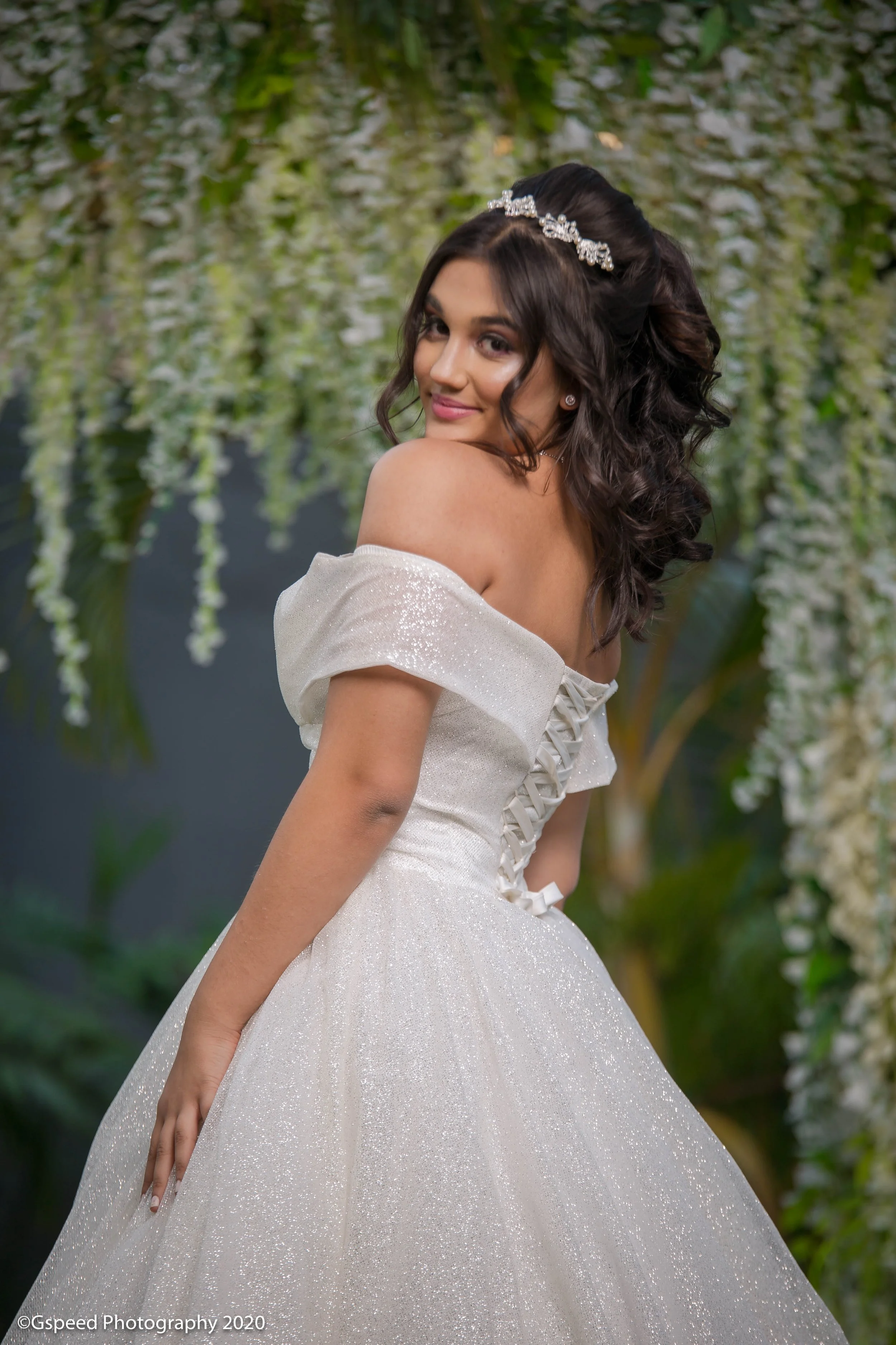 A young woman in a sparkling white off-shoulder dress with lace-up back details posing in front of hanging white flowers and green foliage, smiling at the camera with wavy hair and a tiara in her hair.