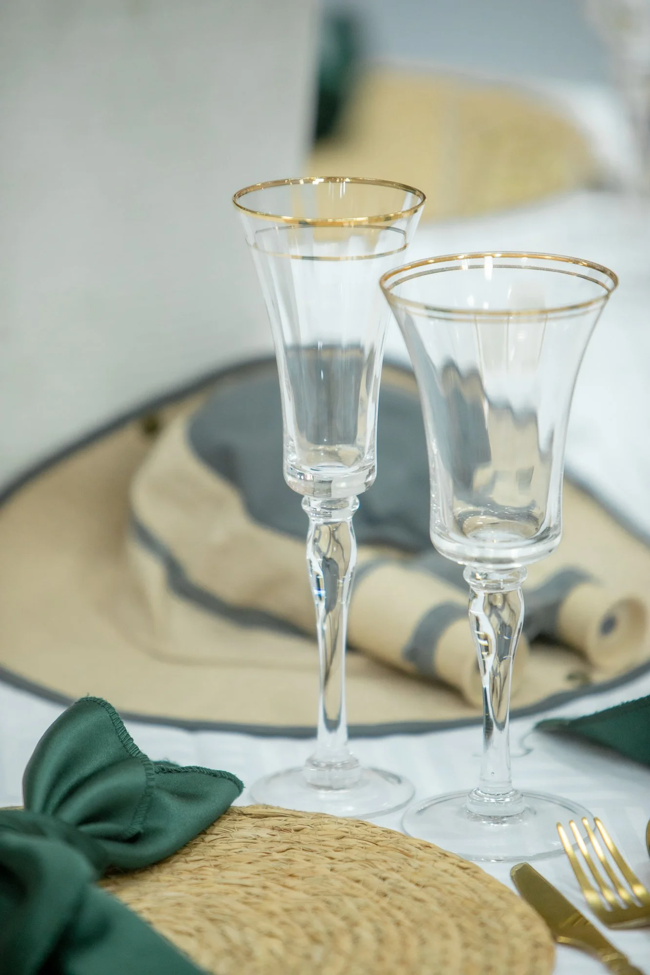 Two clear wine glasses with gold rims on a table. In the background, a tray with a gray napkin and a blurred yellow item. Green cloth and gold cutlery in the foreground.