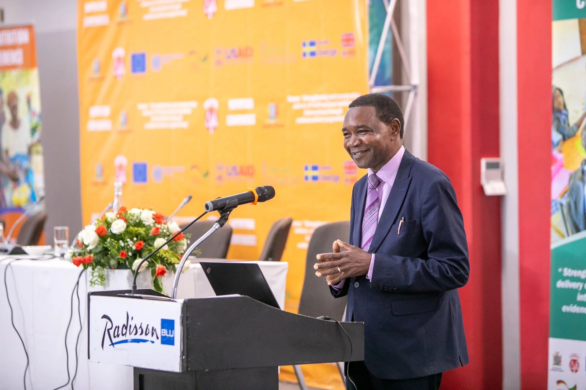 Man in a suit speaking at a conference table with a microphone and laptop, colorful banner in the background, and a flower arrangement on the table.