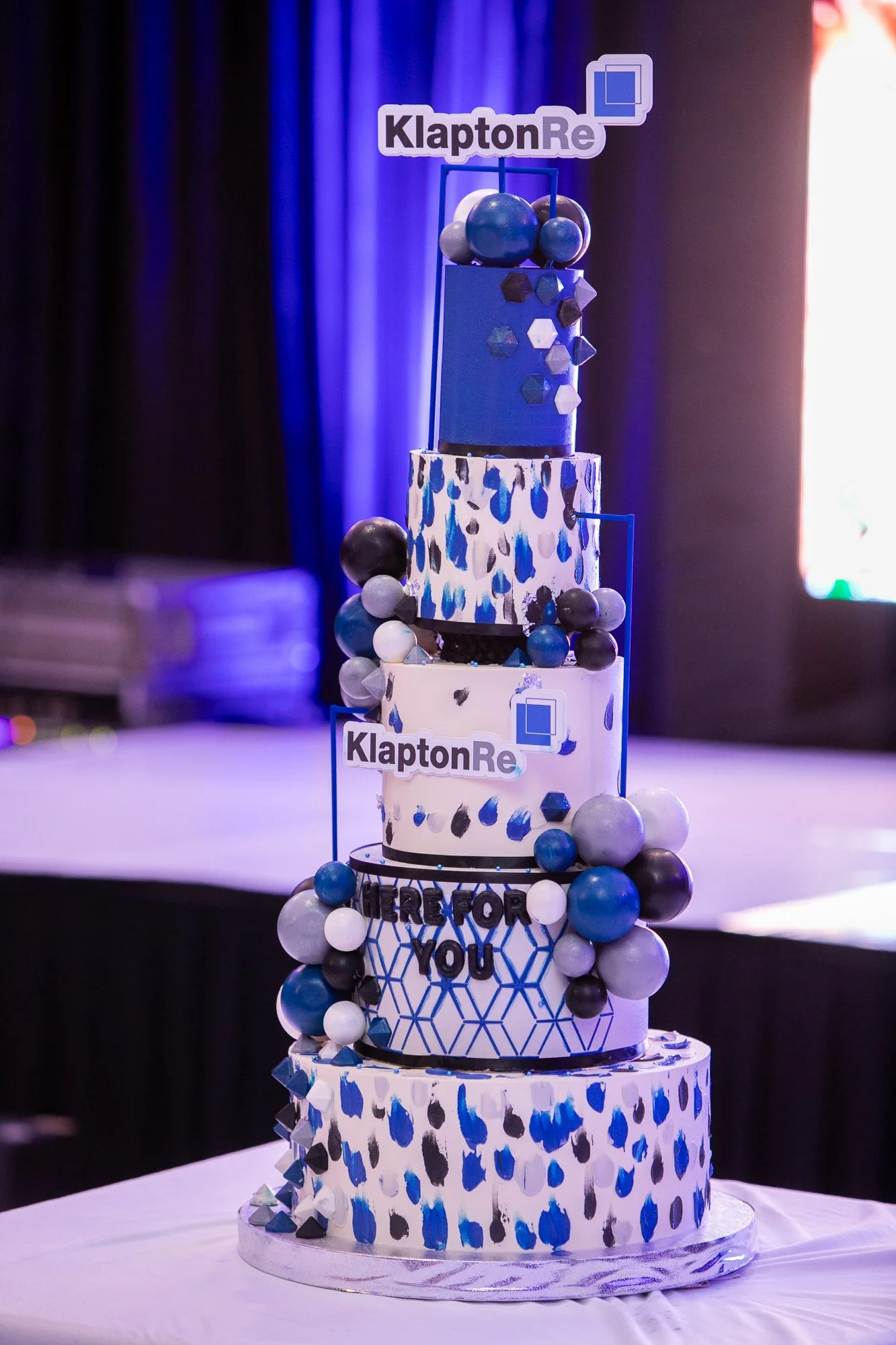 A multi-tiered cake decorated with blue, black, and white elements, featuring the words 'KlaptonRe' and 'Thank you' on the middle tiers, with a blue and white color scheme and geometric patterns, surrounded by spherical decorations.