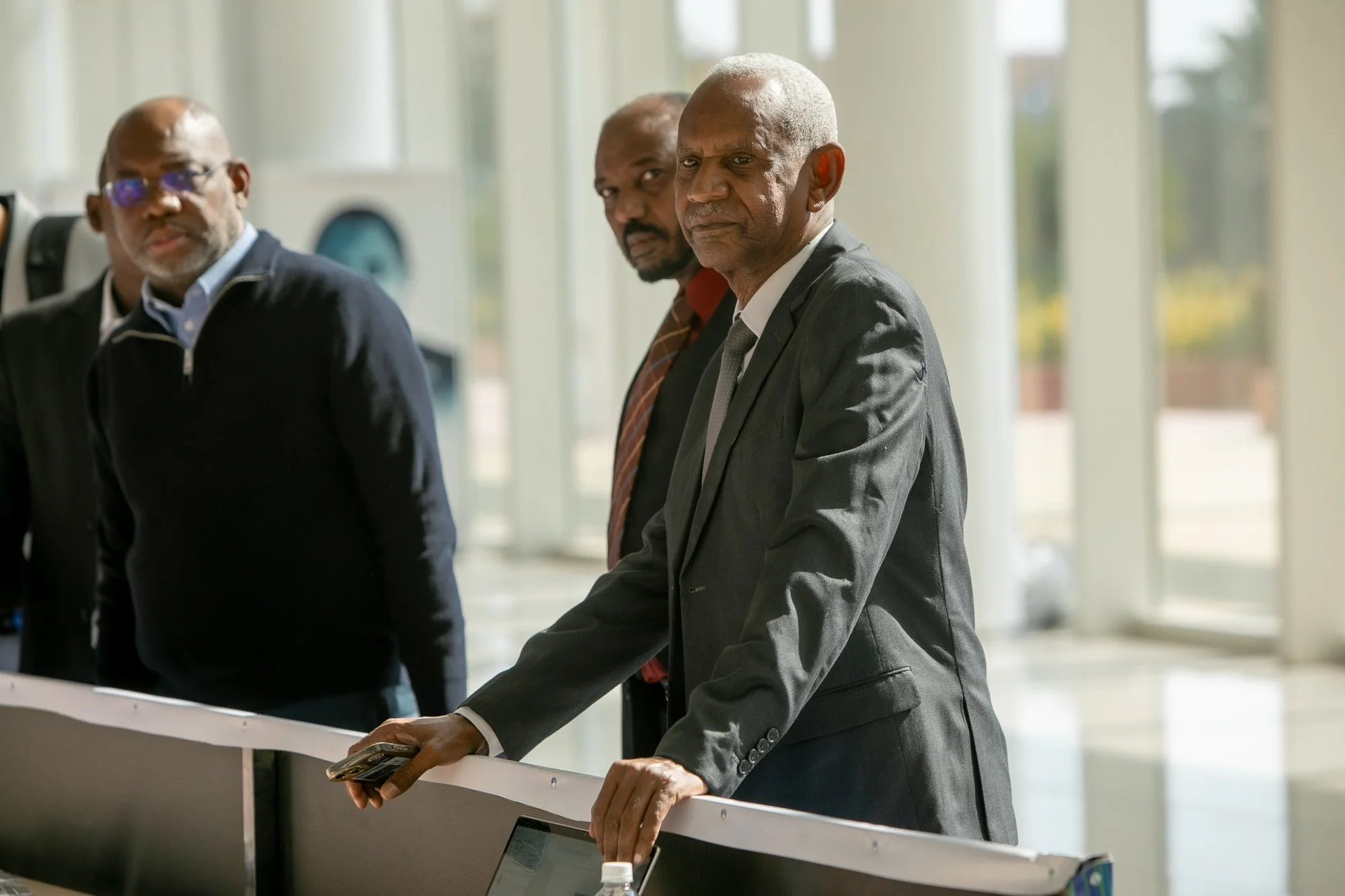 Three elderly men in business attire standing at a counter in a modern building with large windows, one of them holding a smartphone.
