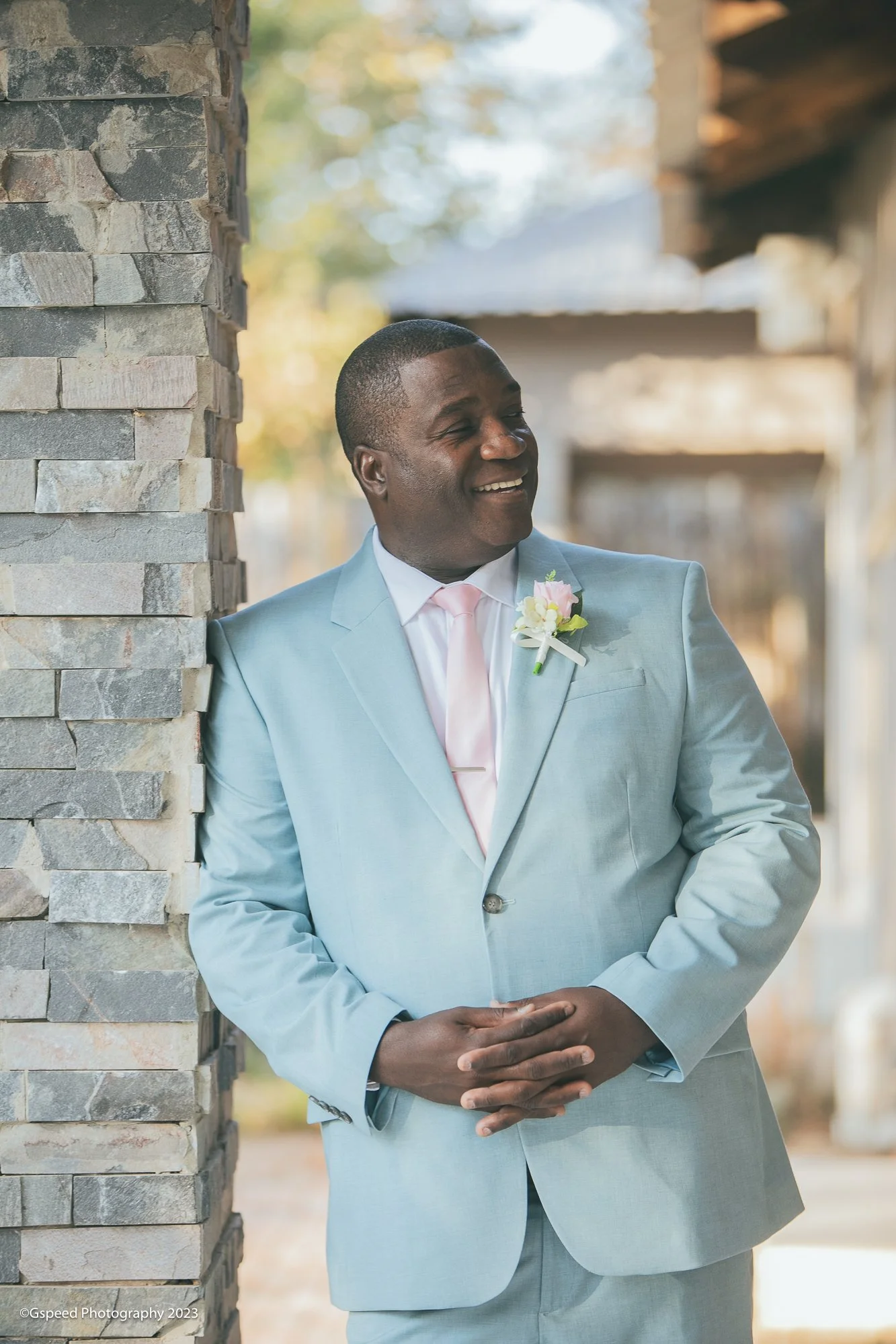 A man in a light blue suit with a pink tie and boutonniere, smiling and leaning against a stone wall outdoors.
