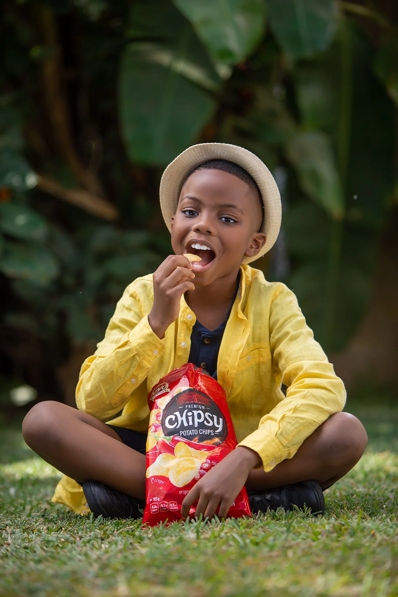 A young boy sitting cross-legged on the grass outdoors, wearing a light beige hat, a yellow jacket, and a dark shirt, eating potato chips from a red bag labeled "Chipsy.