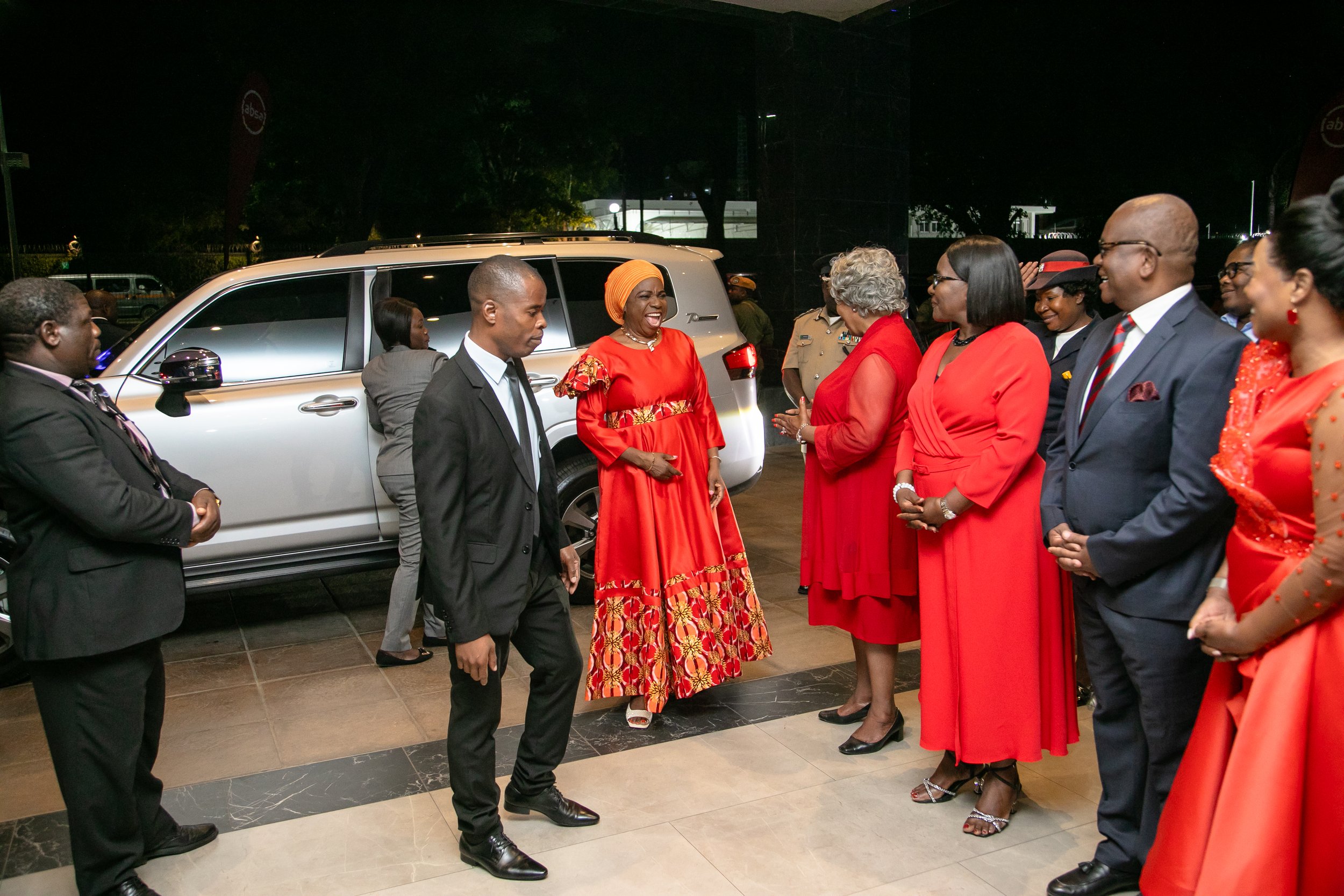 A group of people, dressed in formal and colorful attire, are gathered outdoors at night, engaging in conversation near a white vehicle.