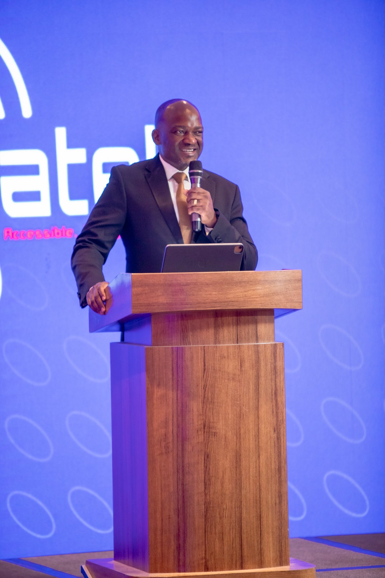 A man in a suit and tie speaking into a microphone at a conference or event, standing behind a wooden podium with a laptop, with a blue background featuring a logo and the words 'Accessible' and 'ate' visible.
