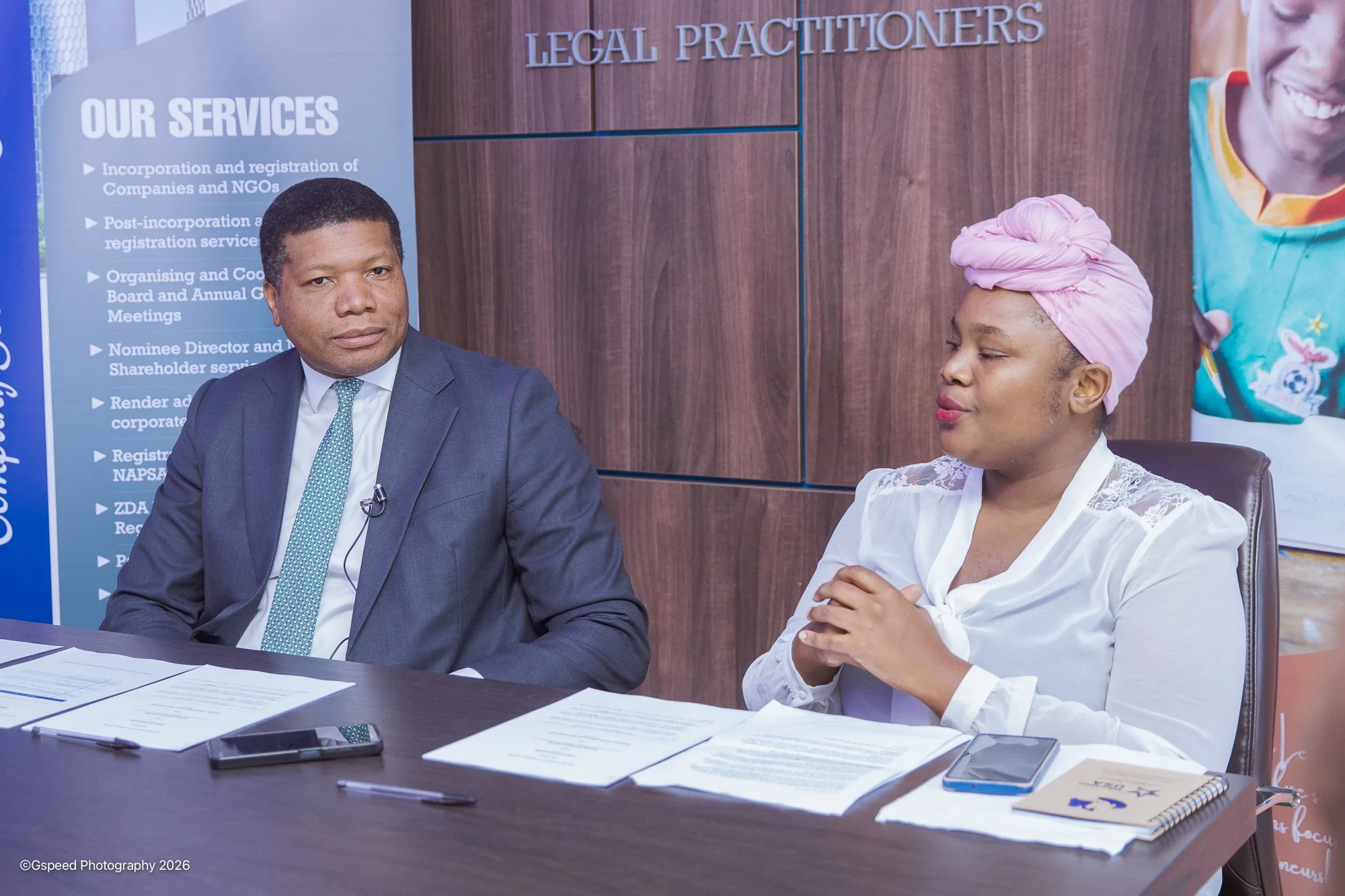 Two people sitting at a desk in a professional setting. The man on the left is wearing a dark suit and tie, looking at the camera. The woman on the right, wearing a white blouse and pink headwrap, has her eyes closed. There are papers, a notebook, an