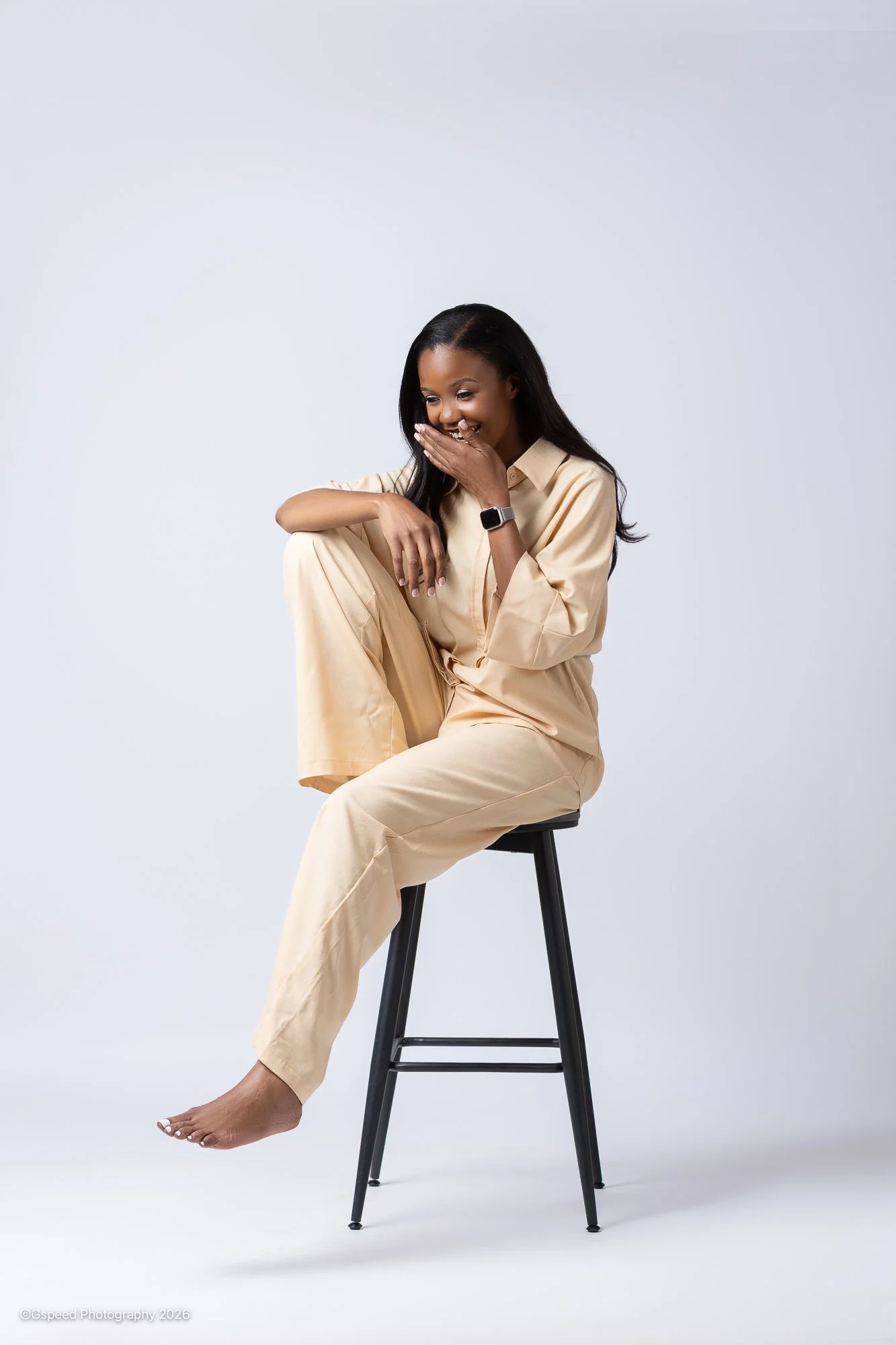 A woman with long black hair wearing beige clothing sitting on a black stool, with one leg crossed over the other, smiling and covering her mouth with her hand, against a plain light gray background.