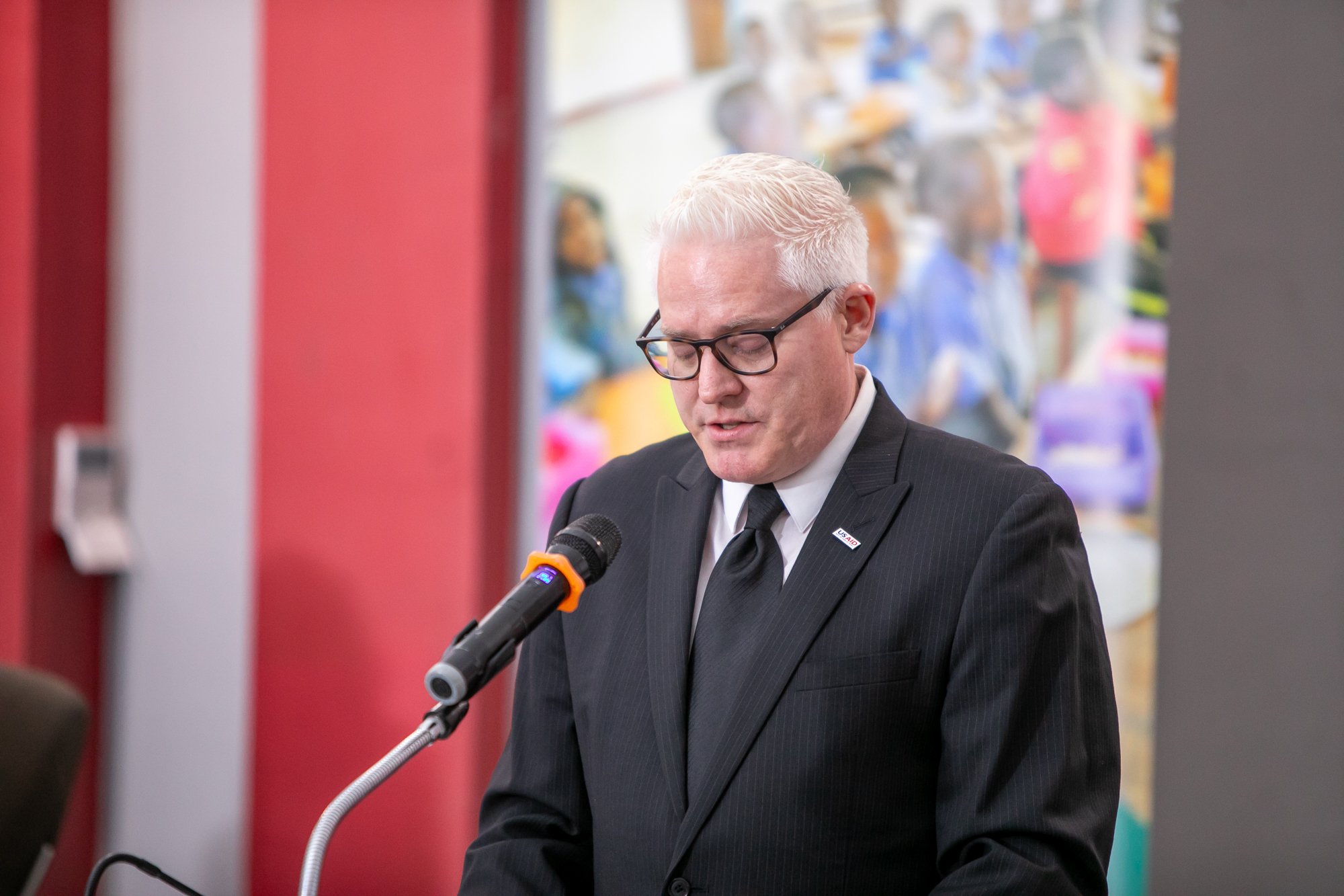 Man in black suit and tie with glasses speaking into a microphone at a public event with people in the background.