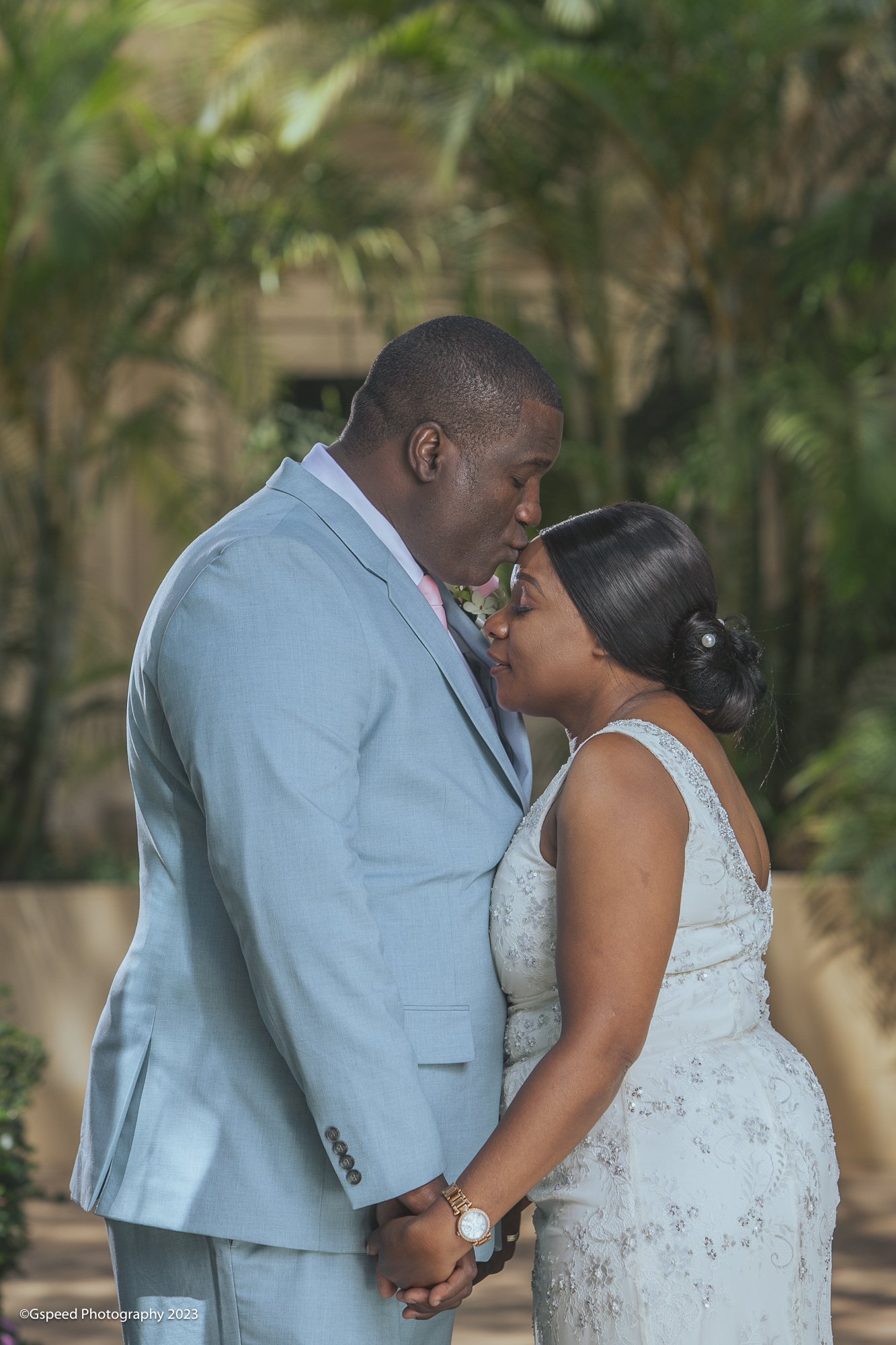 A couple, dressed in wedding attire, holding hands and sharing a tender moment outdoors, with the man kissing the woman's forehead.