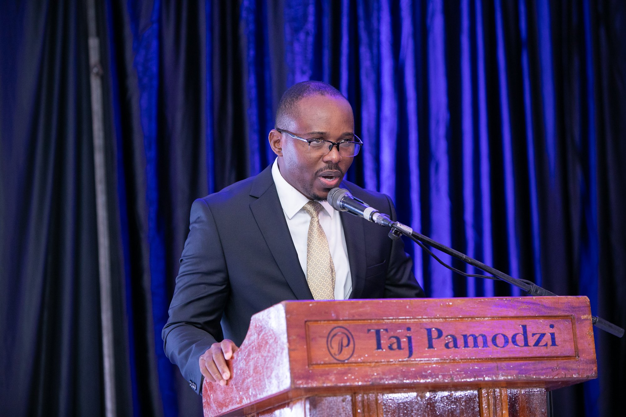 A man in a suit and glasses speaking at a podium with a microphone, holding the edge of the podium, against a blue curtain background.