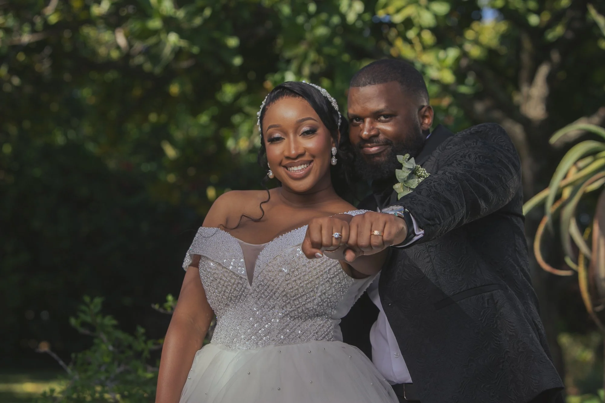 A newlywed African American couple in wedding attire posing outdoors, showing their rings as they joint their hands.