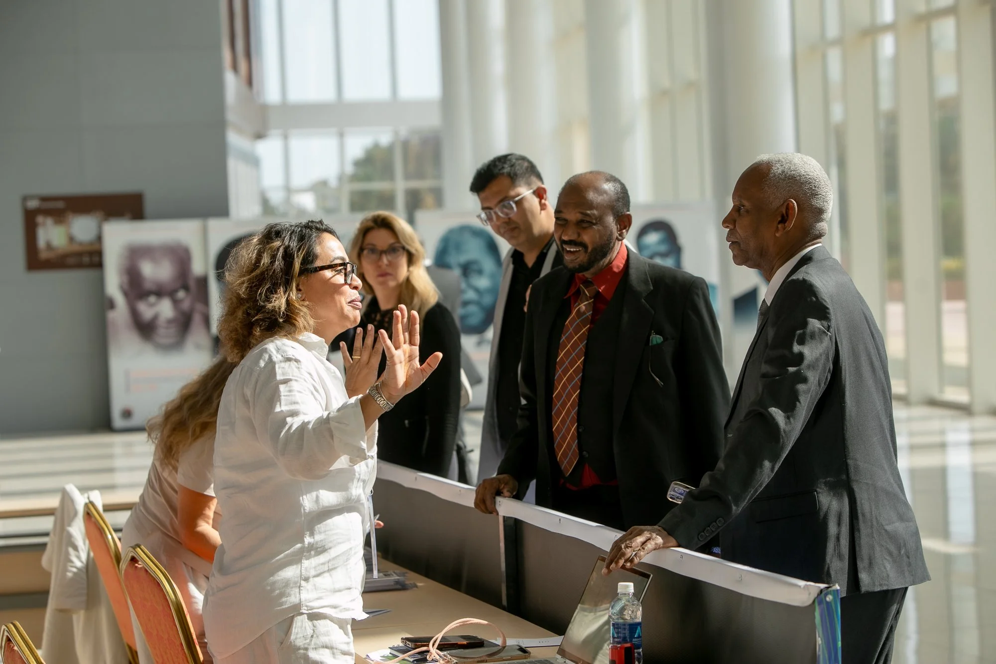 A woman engaging in conversation with four men at a conference or academic exhibition inside a bright, modern building with large windows. The woman is gesturing with her hands, and the men are listening attentively.