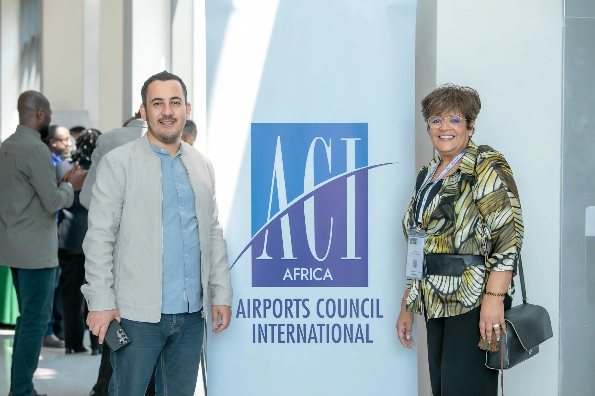 Two individuals standing in front of a sign at an international airports conference, with other people in the background. The man on the left wears a light gray blazer over a blue shirt, holds a phone, and has dark hair and a beard. The woman on the 