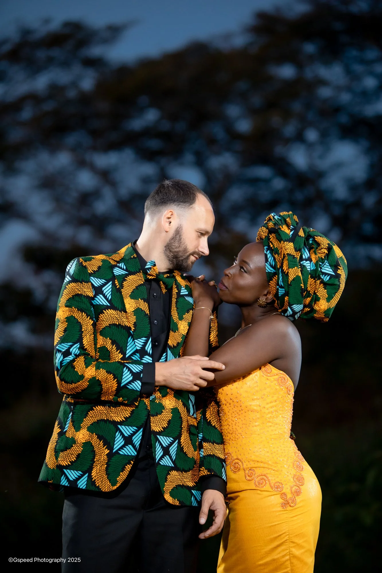 A man and woman stand close together outdoors at dusk, the man is looking down at the woman, and she is gazing back at him. They are both dressed in matching vibrant African print clothing. The woman is wearing a yellow dress with intricate embroider