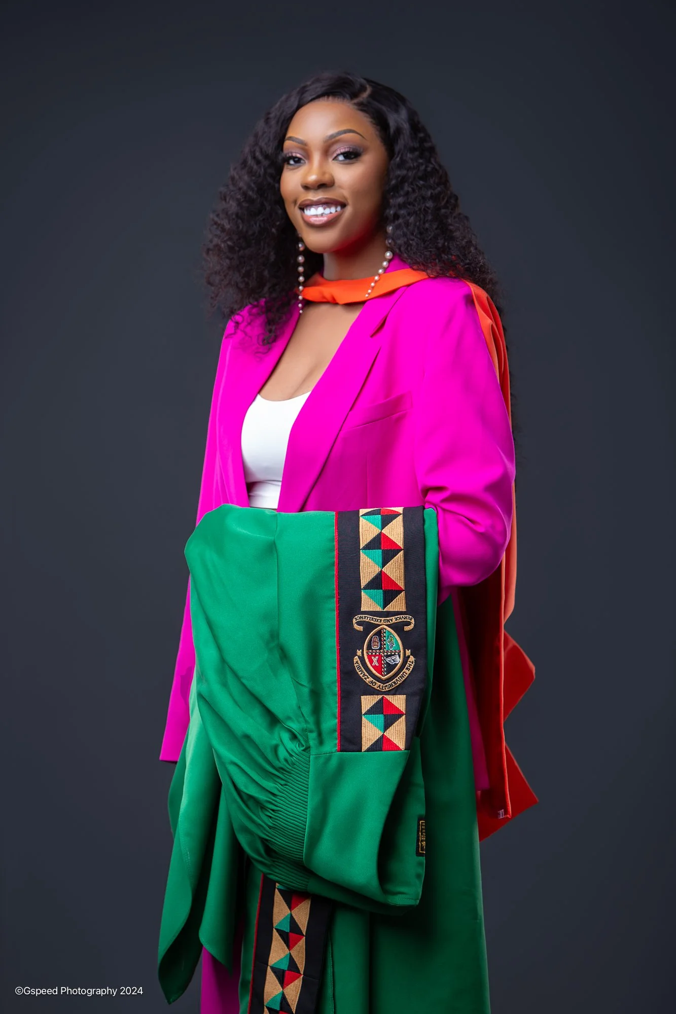 A woman with dark, curly hair wearing a vibrant pink blazer and gold jewelry, holding a green sports jersey with a crest, standing against a dark background, and smiling at the camera.
