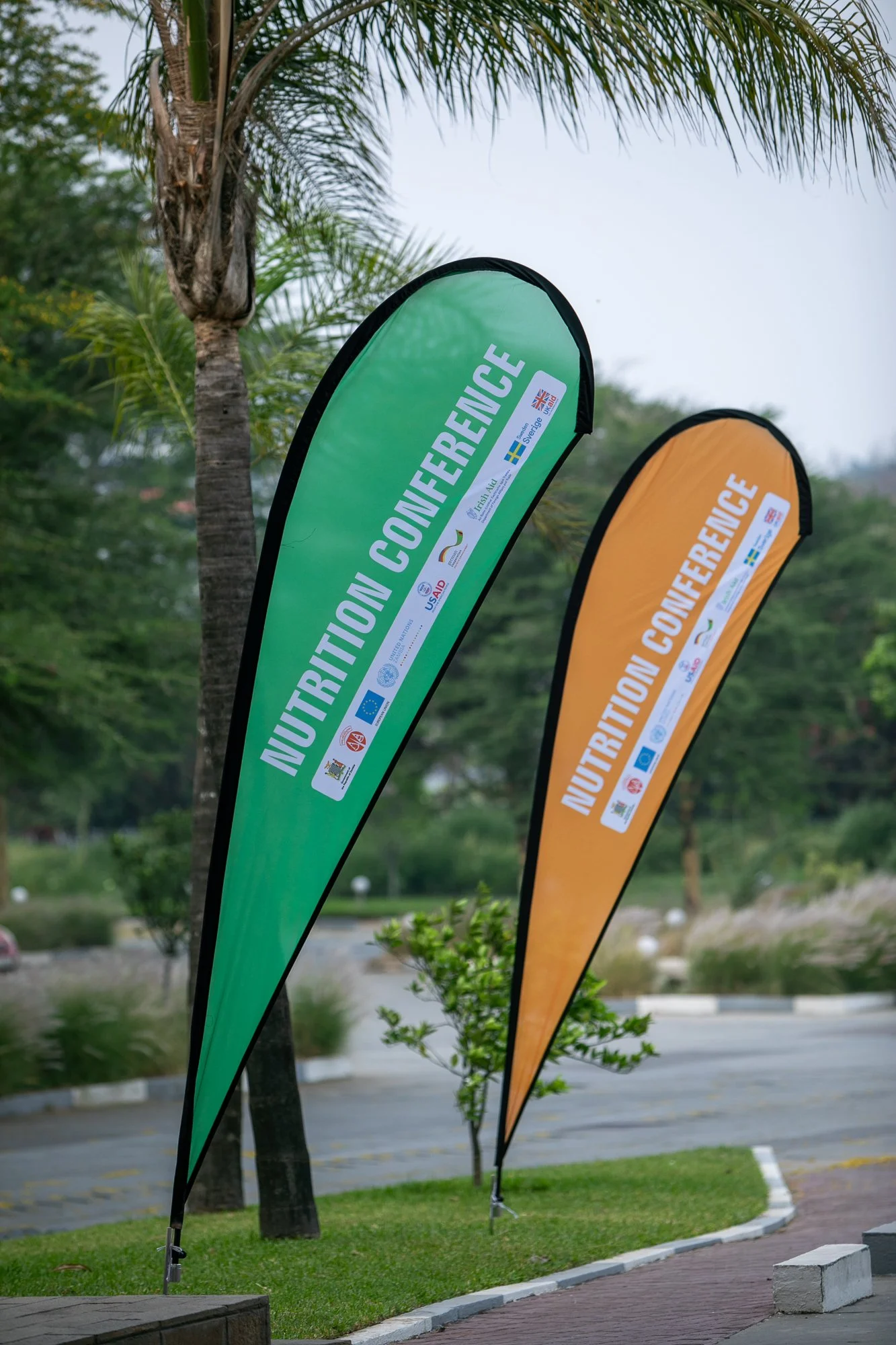 Two vertical flags on a sidewalk with palm trees in the background. The green flag reads 'Nutrition Conference' and has logos of various organizations, while the orange flag has similar text and logos.