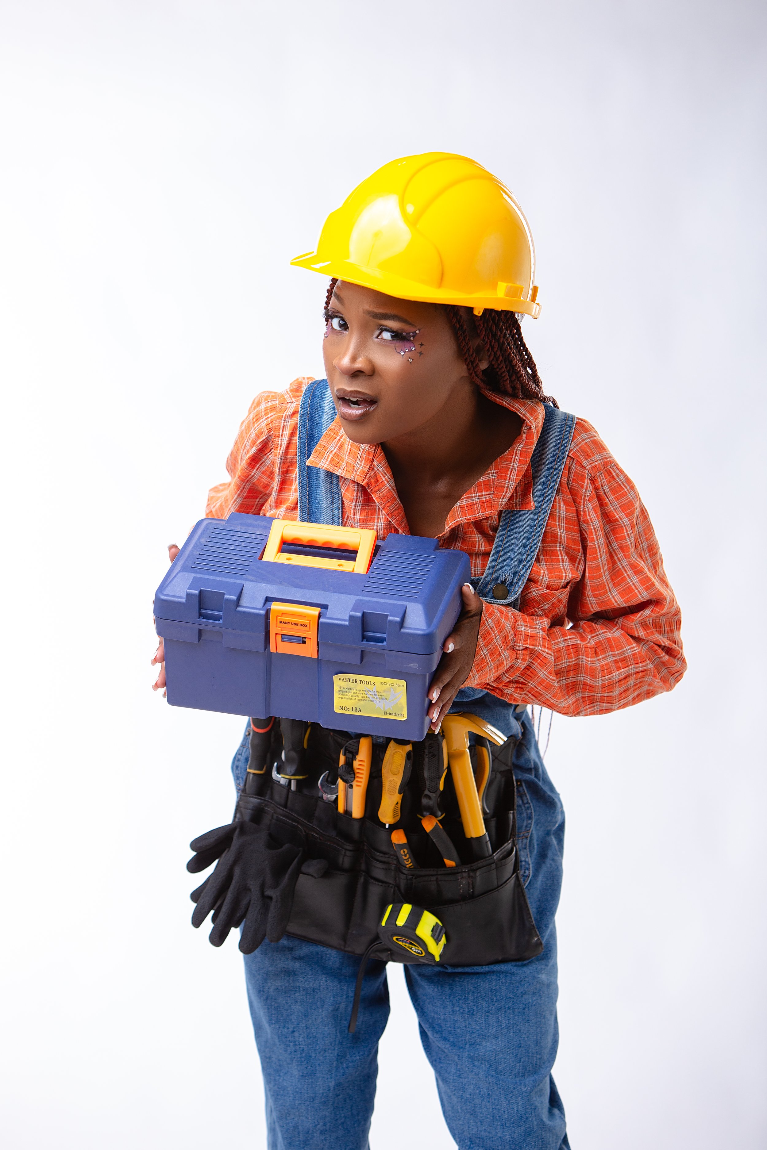 A woman wearing a yellow construction hard hat, makeup with decorative stars, an orange plaid shirt, and blue jeans, holding a blue toolbox with tools and standing against a plain white background.