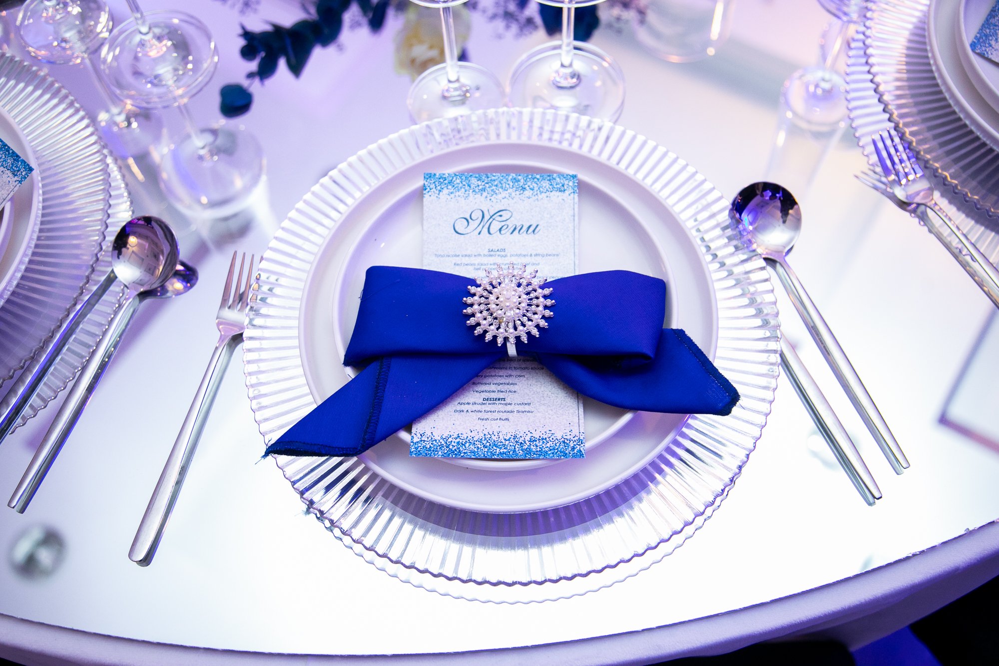 An elegant table setting with a white plate, a blue napkin pinned with a decorative brooch, and a menu card. Surrounding the plate are wine glasses, silverware, and additional plates, all arranged on a white tablecloth with a textured rim.