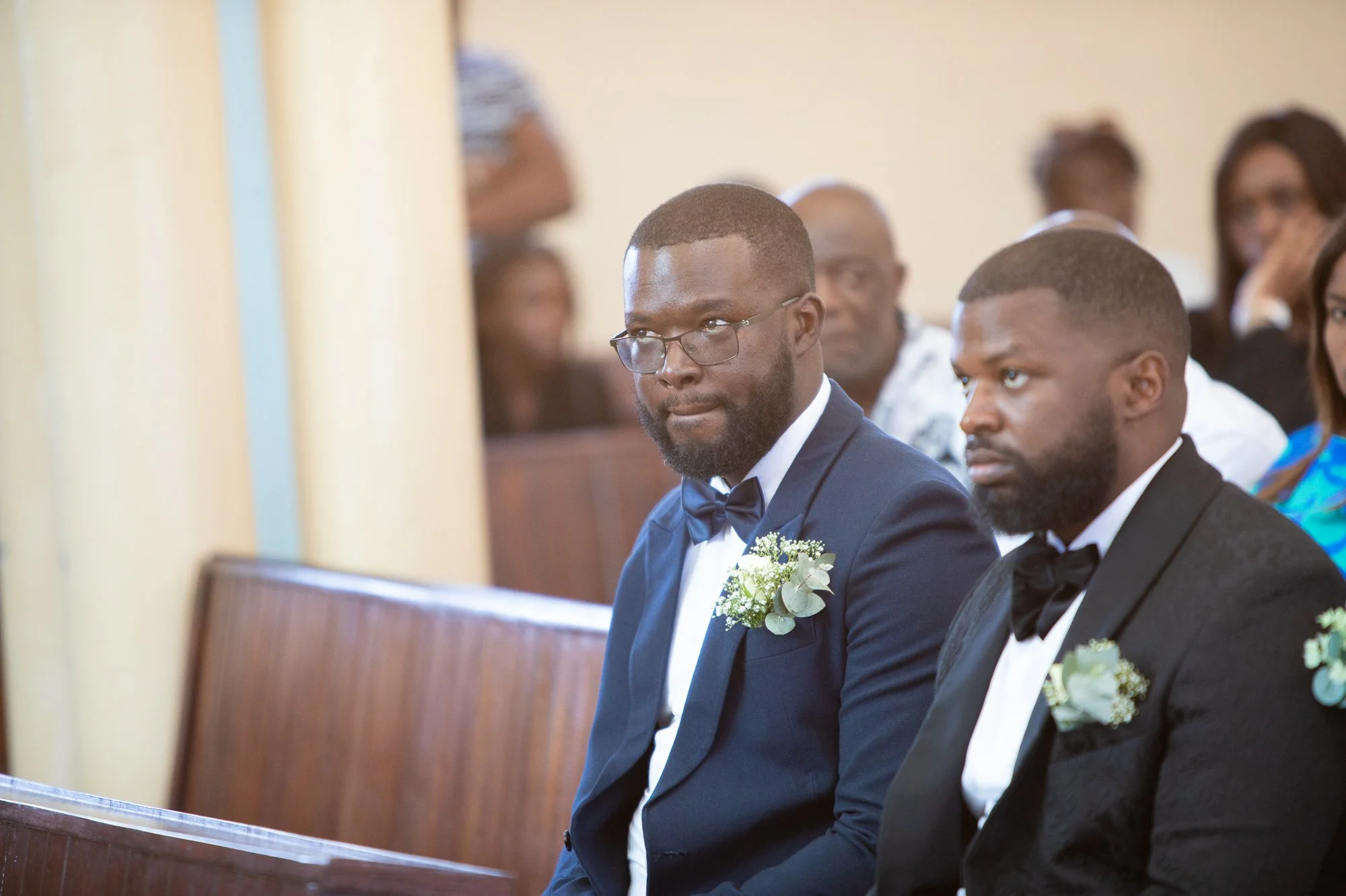A groom in a tuxedo with a boutonniere is sitting in a church alongside the best man and other groomsmen, attending a wedding ceremony.