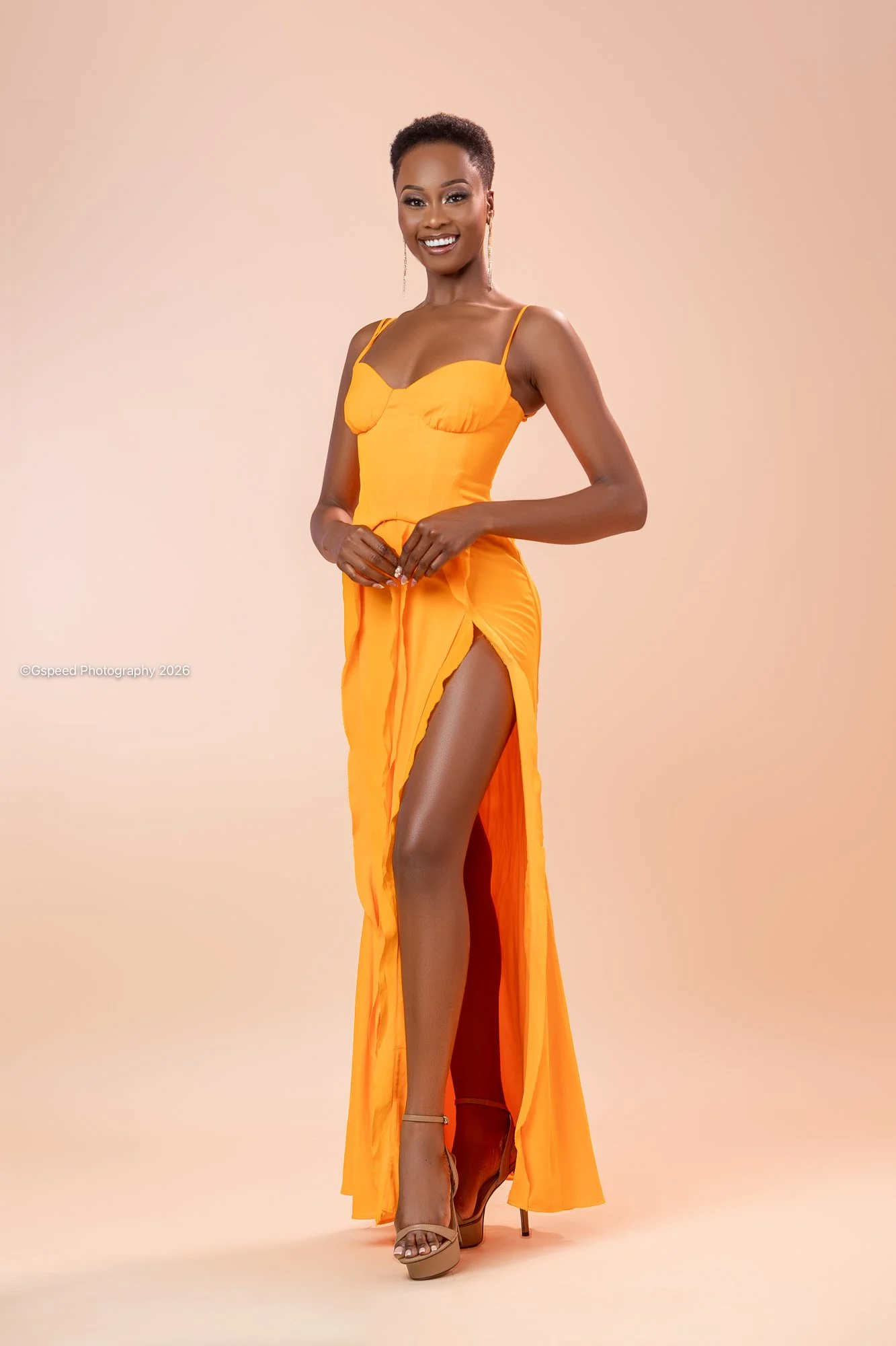 A woman modeling an orange dress with a high slit, paired with high heels, standing against a neutral background, smiling and looking at the camera.